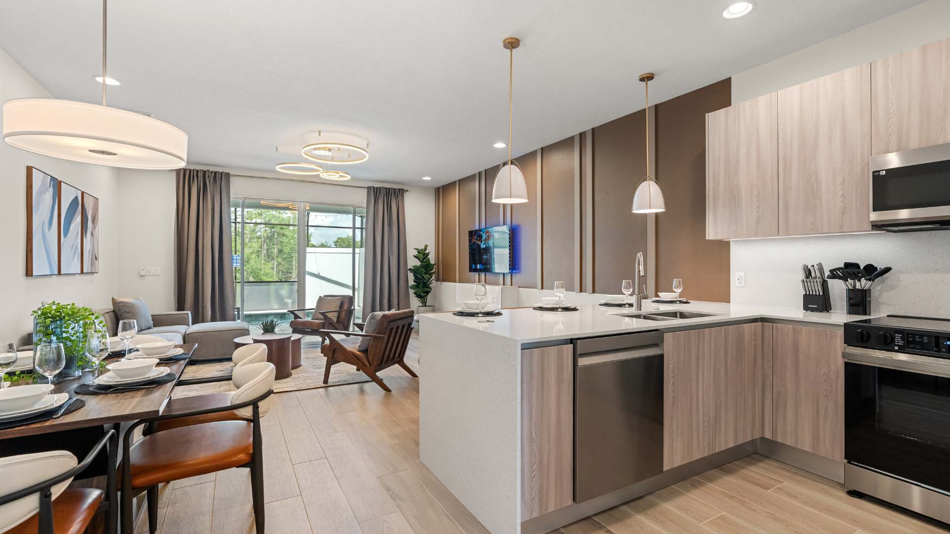 Open-concept living space featuring a modern kitchen island with seating, a dining area, and a living room with a gray sectional sofa and large windows. A decorative brown panel wall adds a stylish accent.