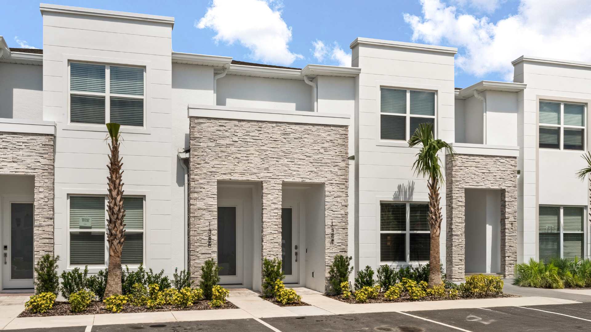 Modern white townhouse exterior with light-colored stone accents around the entrances and large windows. Palm trees and low-lying shrubs decorate the front, with paved parking visible.