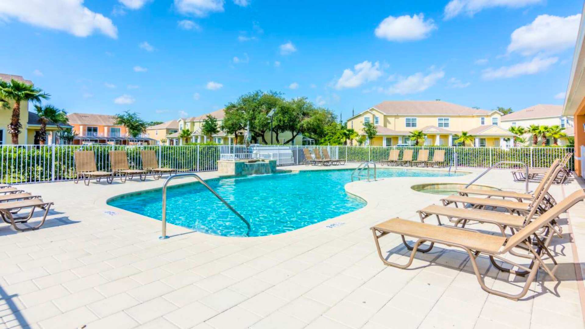A large, irregularly shaped communal swimming pool with clear blue water, surrounded by light-colored pavers and numerous lounge chairs, set against a backdrop of yellow and orange multi-story buildings under a bright blue sky.