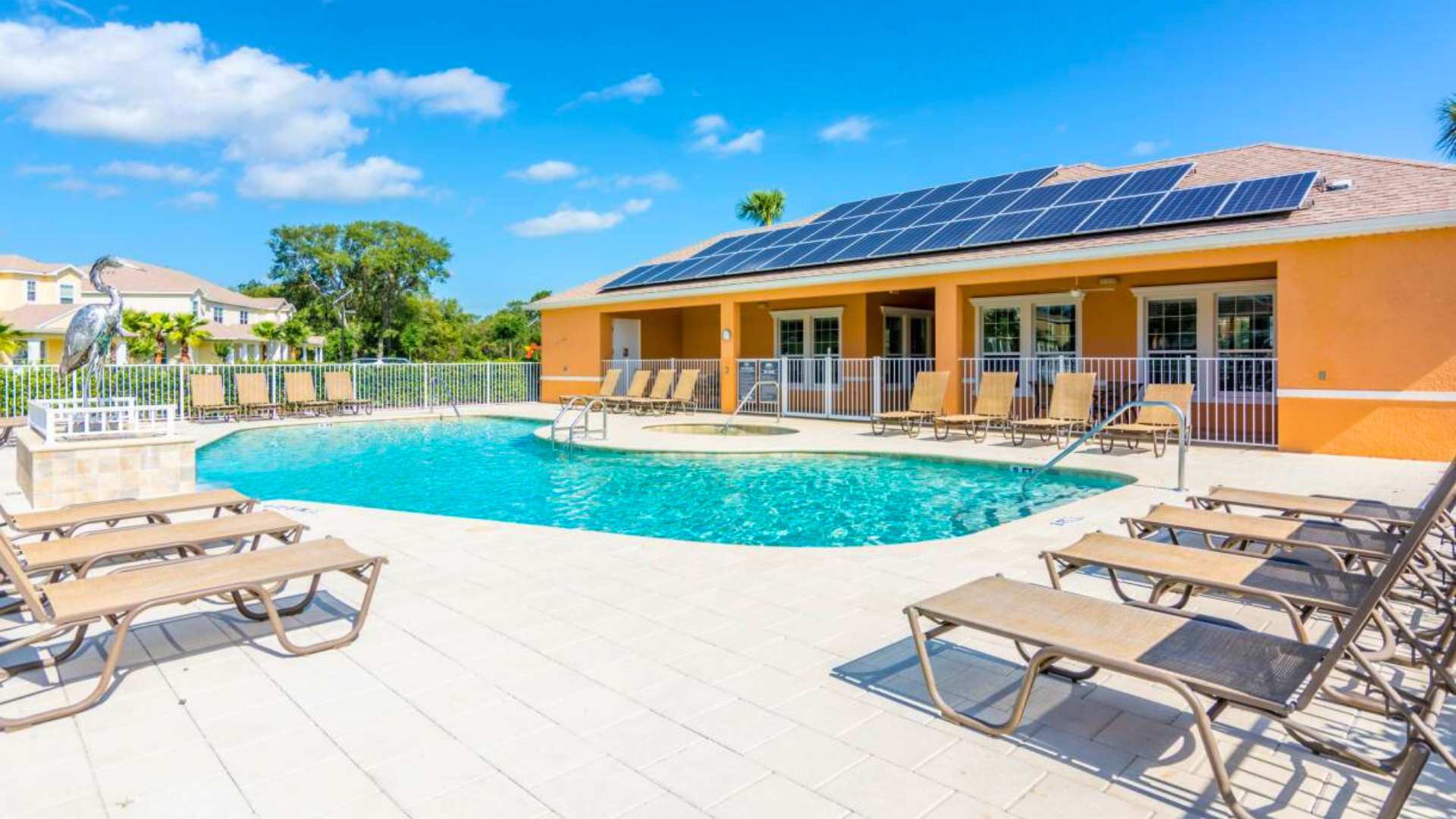 A vibrant orange clubhouse building with solar panels on its roof overlooks a communal swimming pool and hot tub area, surrounded by lounge chairs and a white fence, under a bright blue sky.