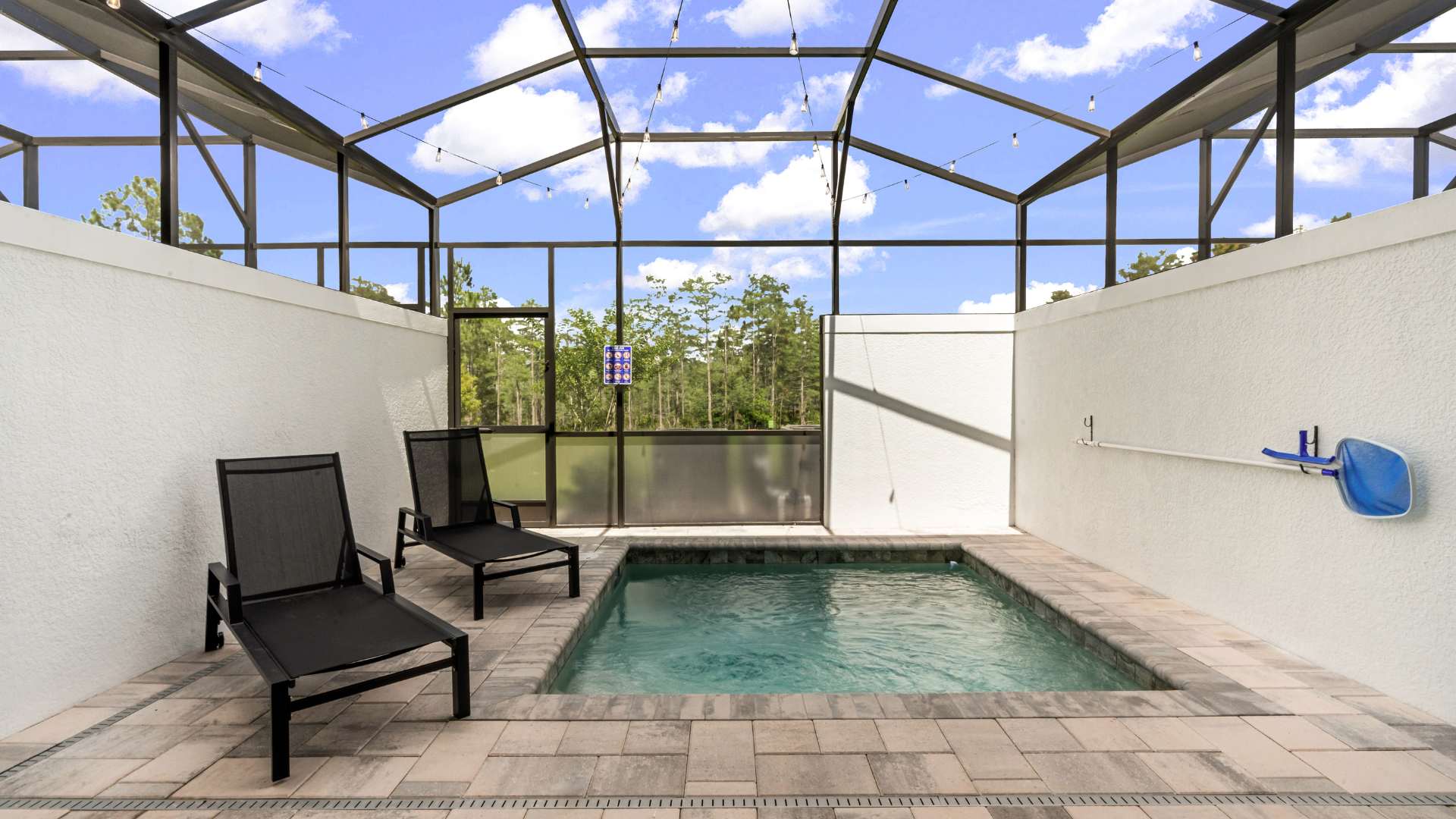 Private screened-in plunge pool area featuring two black lounge chairs on light pavers, with a view of trees outside the enclosure. The pool has a small waterfall feature, and string lights are strung across the ceiling.