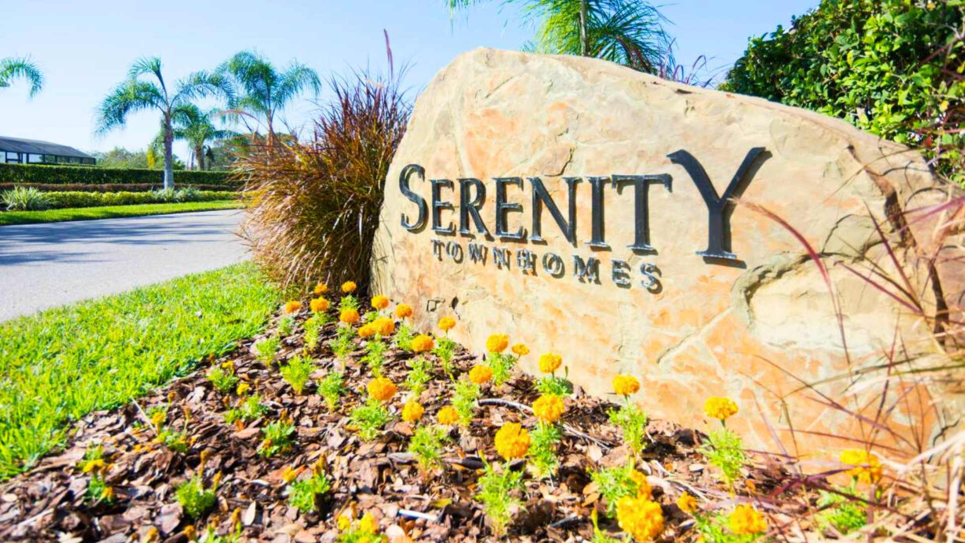 Large stone sign for 'Serenity Townhomes' surrounded by green grass and yellow flowers. The sign is carved with the community name, indicating the property's location.