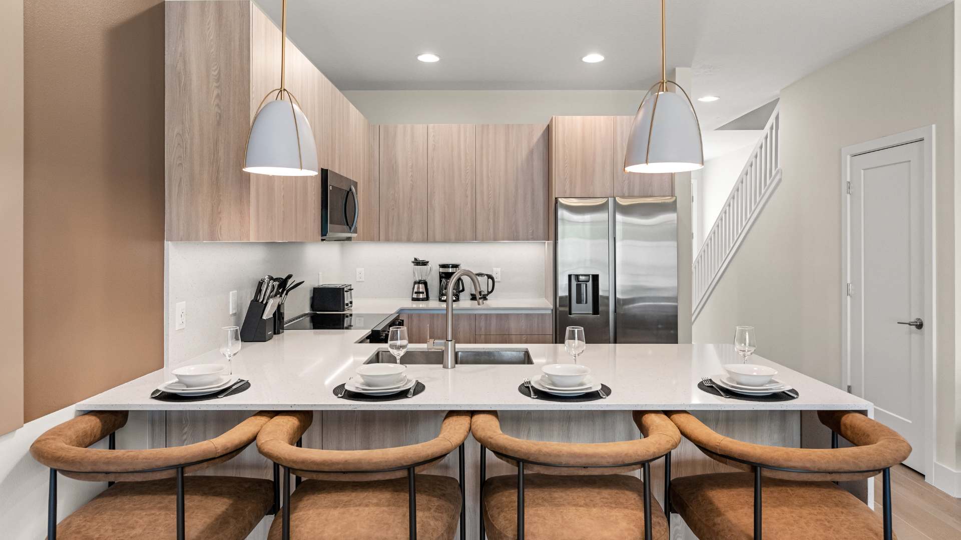 Bright kitchen with light wood cabinets, white quartz countertops, and a large island with a double sink and four brown leather bar stools. A stainless steel refrigerator is visible, and a staircase is in the background.