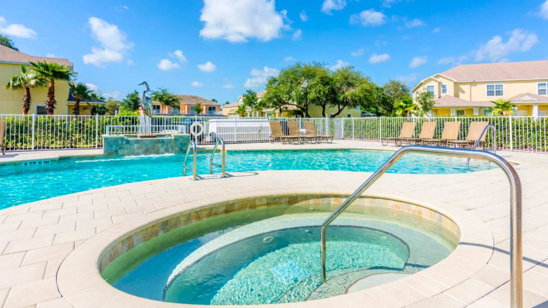 A sparkling hot tub with steps and a metal handrail, adjacent to a communal swimming pool, both filled with clear blue water and surrounded by light-colored pavers and stone accents, under a bright blue sky.