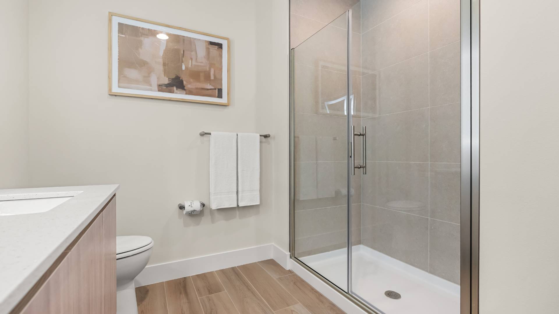 Contemporary bathroom with a glass-enclosed walk-in shower featuring light gray tiled walls and a white shower pan. A toilet is visible next to the vanity, and a framed abstract artwork hangs on the wall.