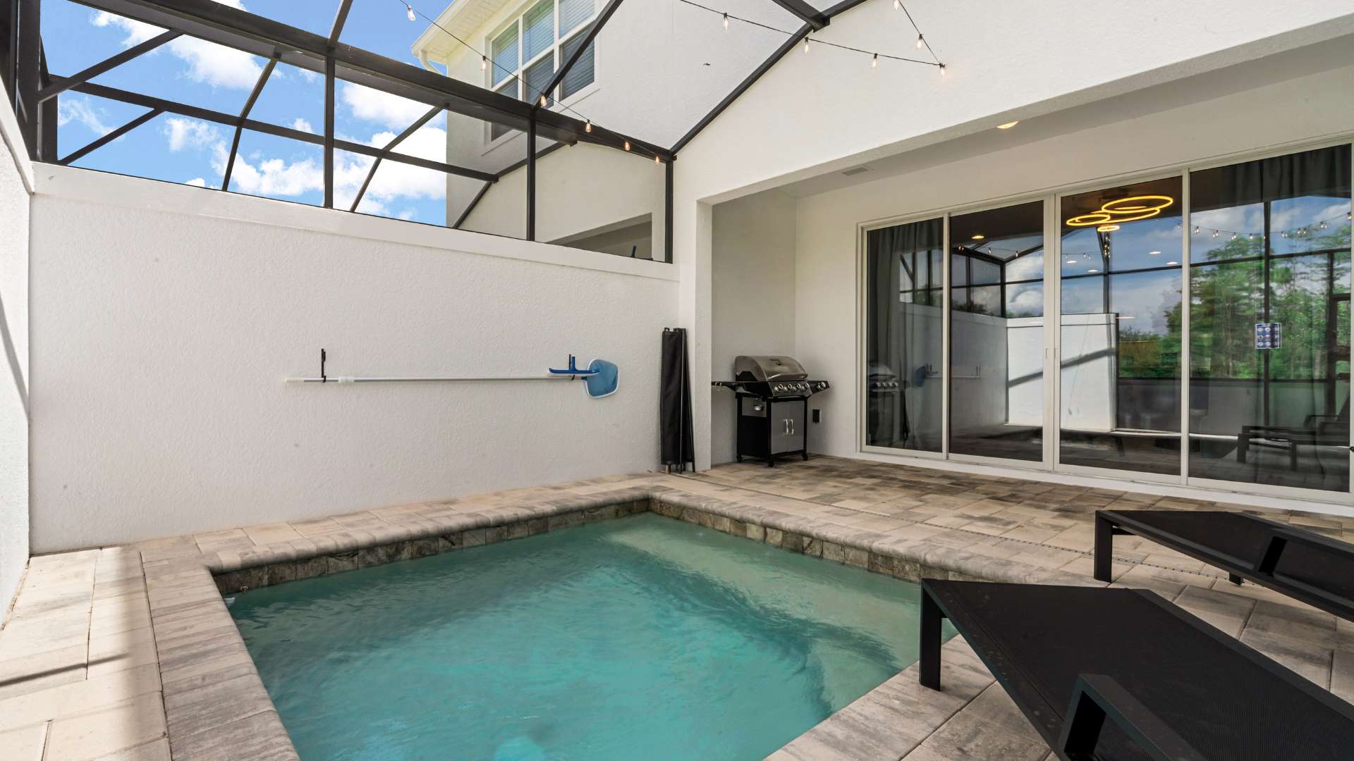Screened-in private plunge pool area with light-colored pavers, a built-in BBQ grill, and string lights overhead. The pool is surrounded by white walls, and a sliding glass door leads into the house.