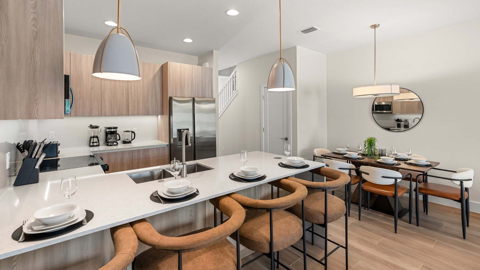 Modern kitchen with light wood cabinetry, white quartz countertops, and a large island featuring a double sink and four brown leather bar stools. A dining area with a wooden table and chairs is visible in the background.