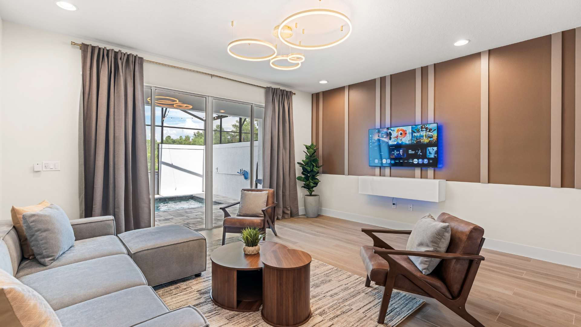 Modern living room with light wood floors, a gray sectional sofa, and two brown leather armchairs. A large flat-screen TV is mounted on a textured accent wall, and sliding glass doors lead to the private pool area.