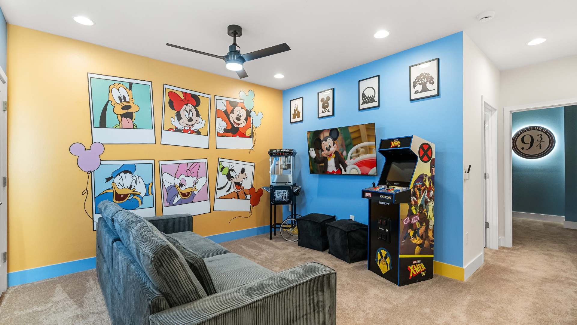 Vibrant game room with a blue and yellow accent wall featuring large Disney character polaroid-style artwork. The room includes a gray sofa, a flat-screen TV, a popcorn machine, and an arcade game machine.