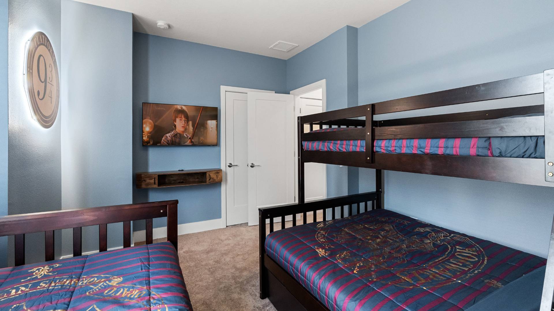 Another view of the themed children's bedroom, showcasing a wall-mounted TV playing a movie and a prominent 'Platform 9 3/4' sign with backlighting. The room has light blue walls and dark wood furniture.