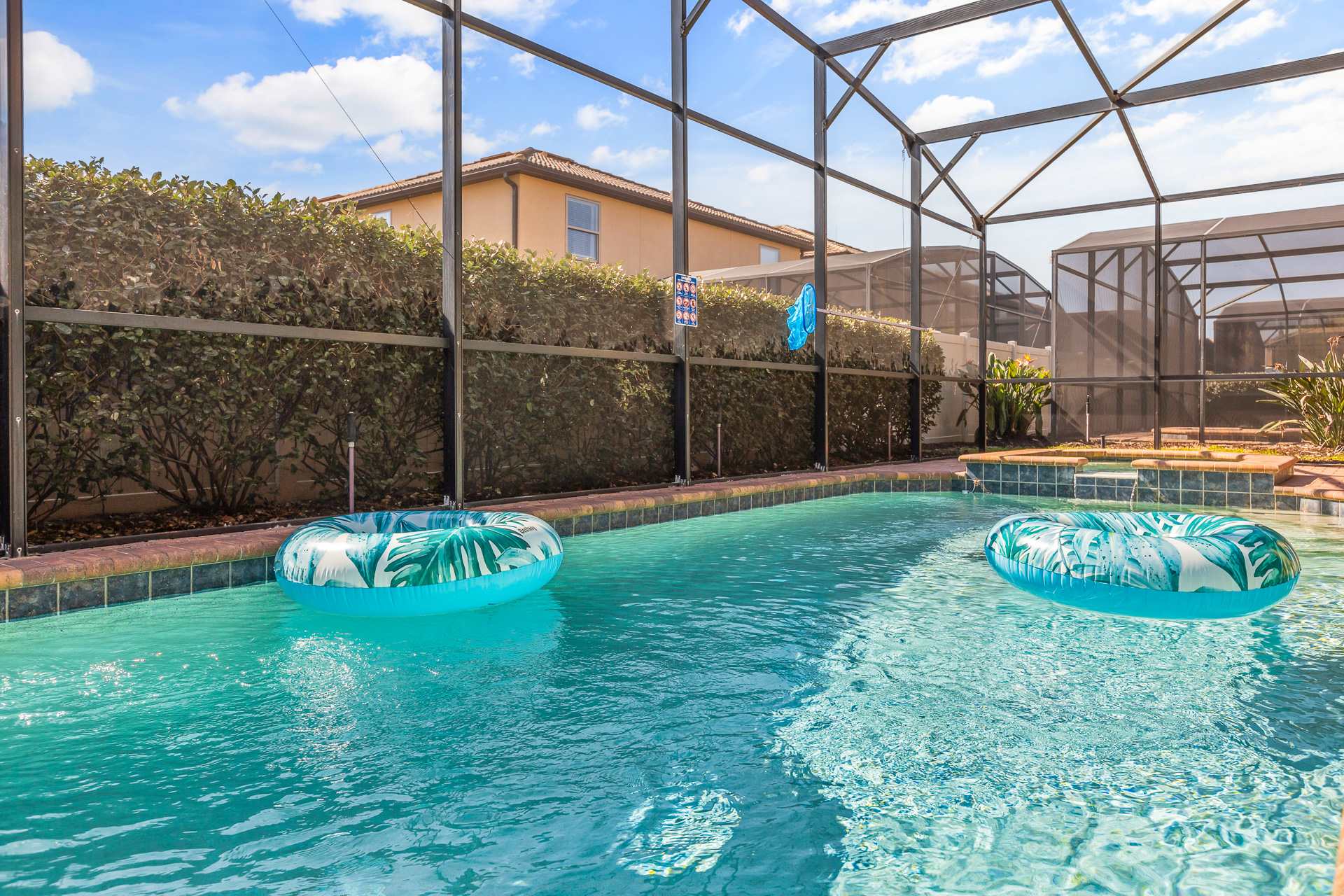 A close-up view of the private screened pool with two tropical-patterned inflatable floats, showing the surrounding hedge and the neighboring house in the background.