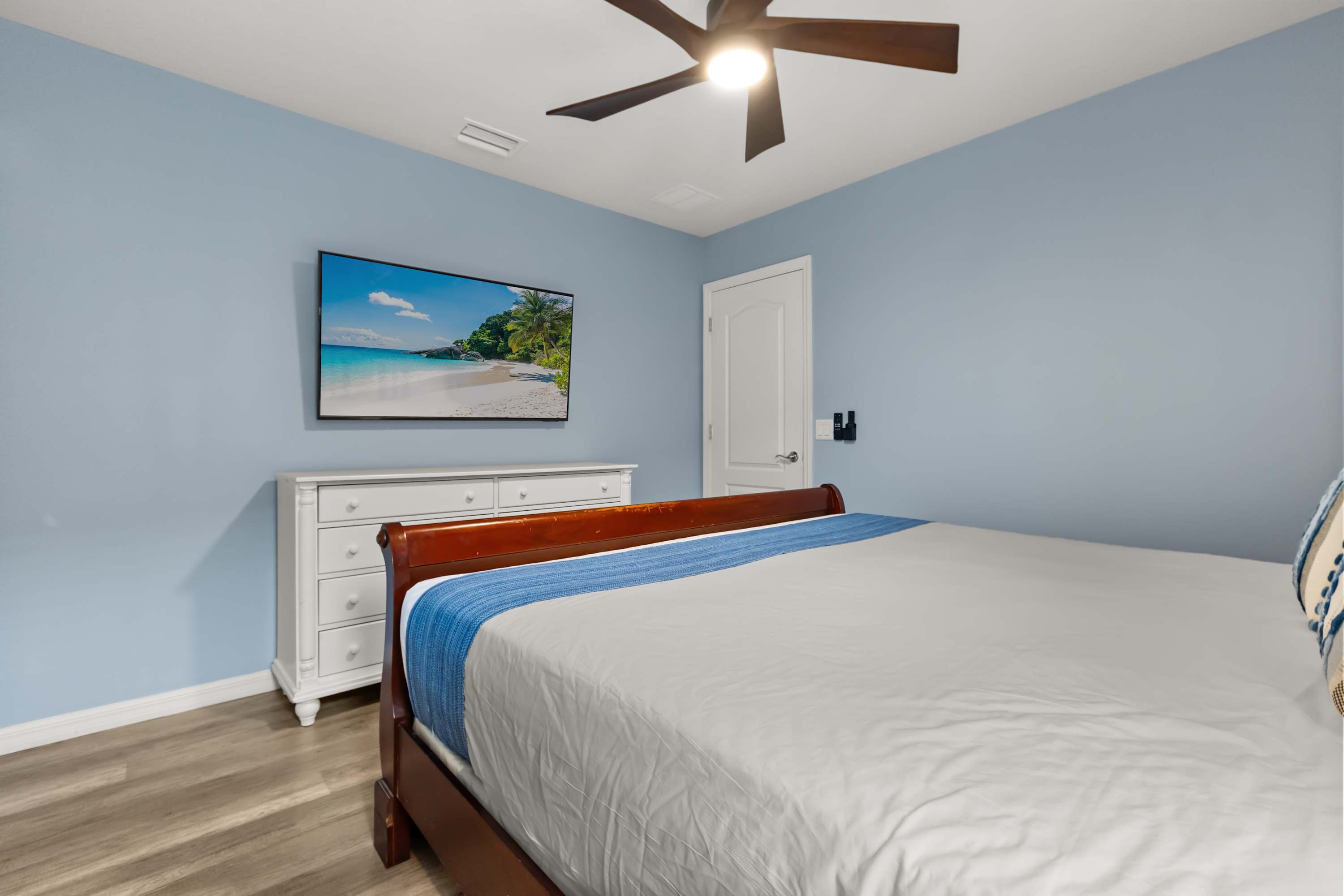 Another angle of the light blue bedroom, showing a white dresser with a wall-mounted TV displaying a beach scene. The large wooden bed with white bedding and a blue throw is also visible.