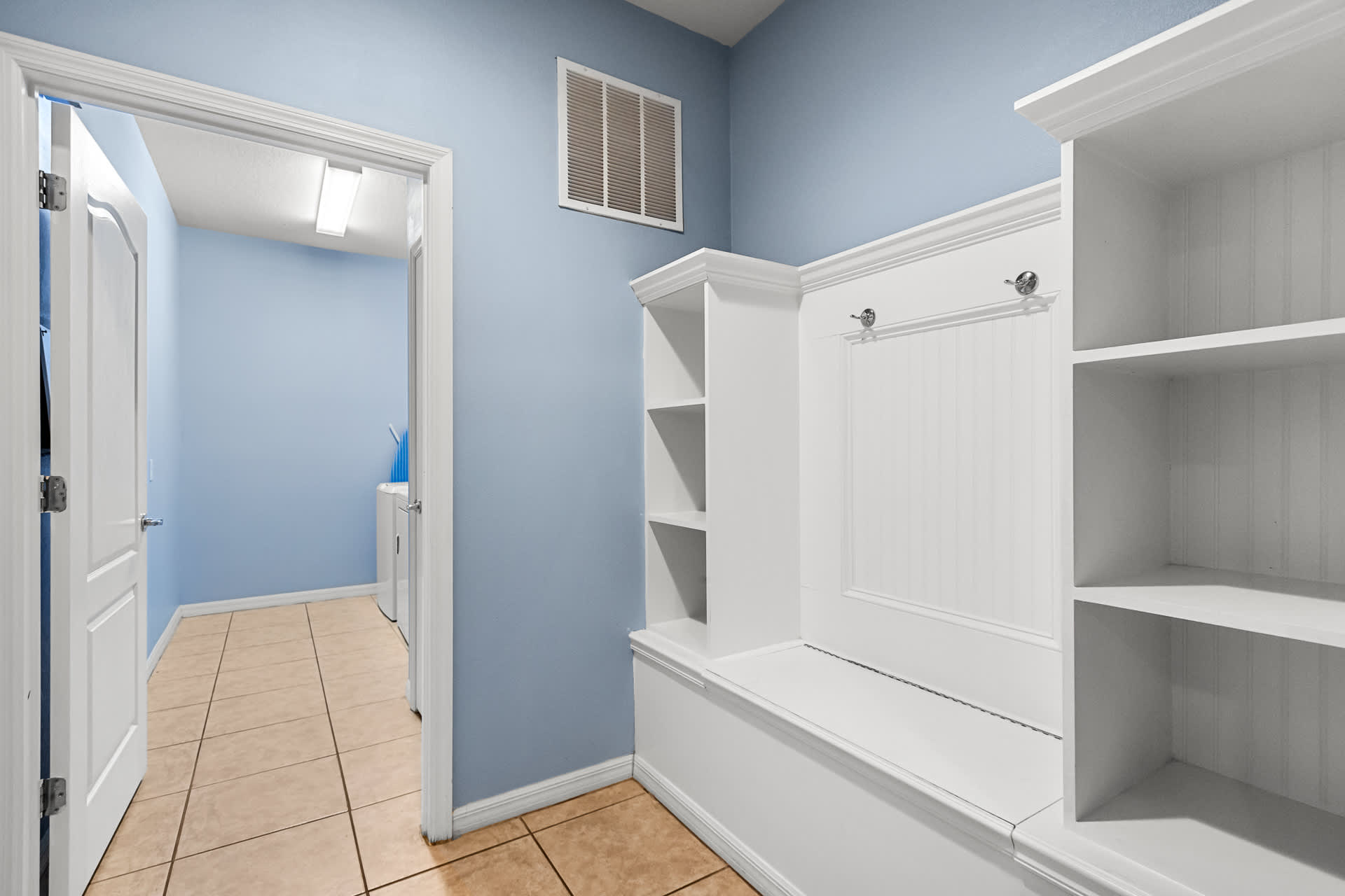 A light blue mudroom features a white built-in bench with cubbies and hooks, providing storage. An open doorway reveals a laundry room with a washer and dryer.