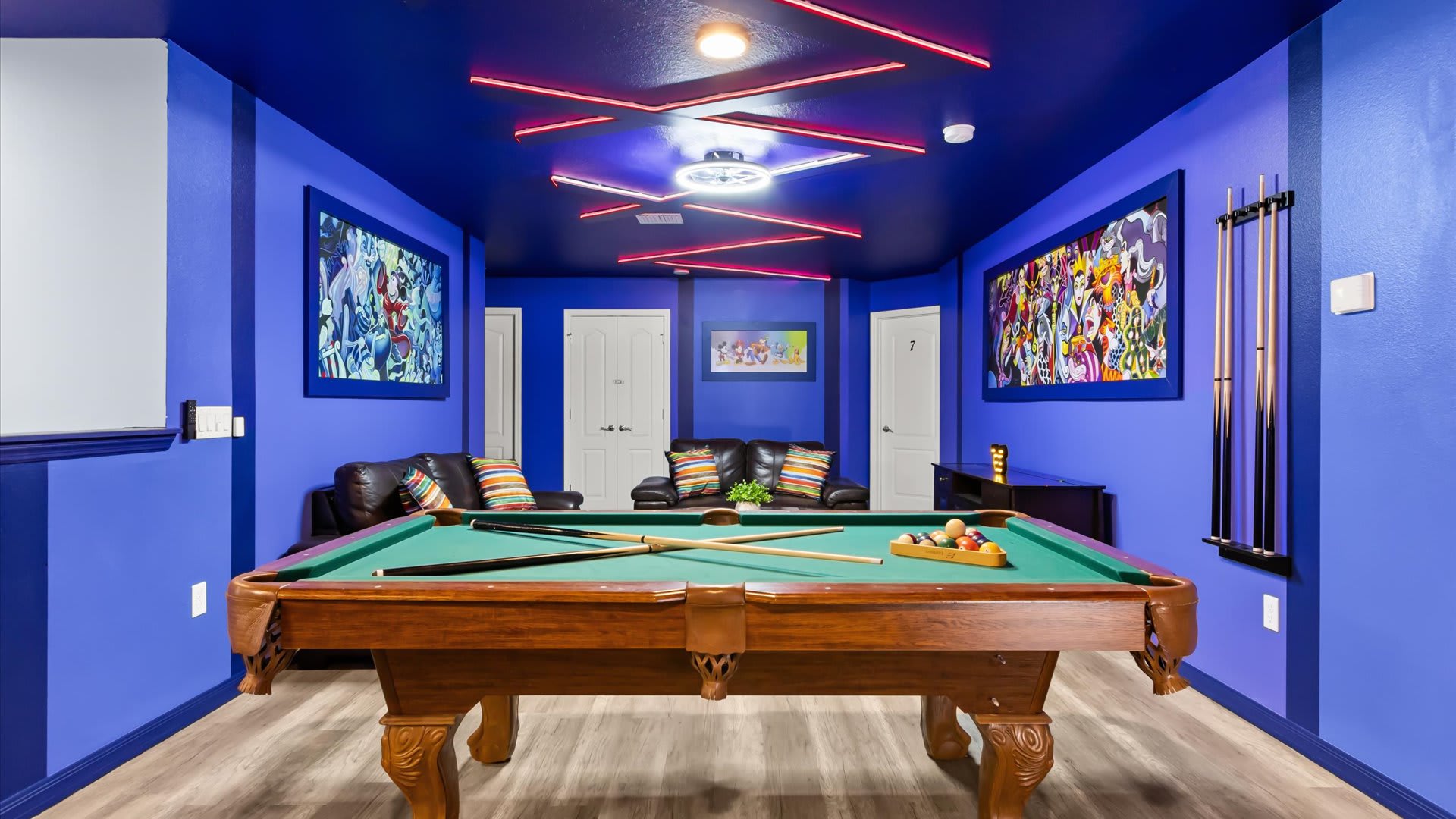 A vibrant game room featuring a wooden pool table with green felt, surrounded by blue walls with colorful framed artwork, and a seating area with dark leather sofas.