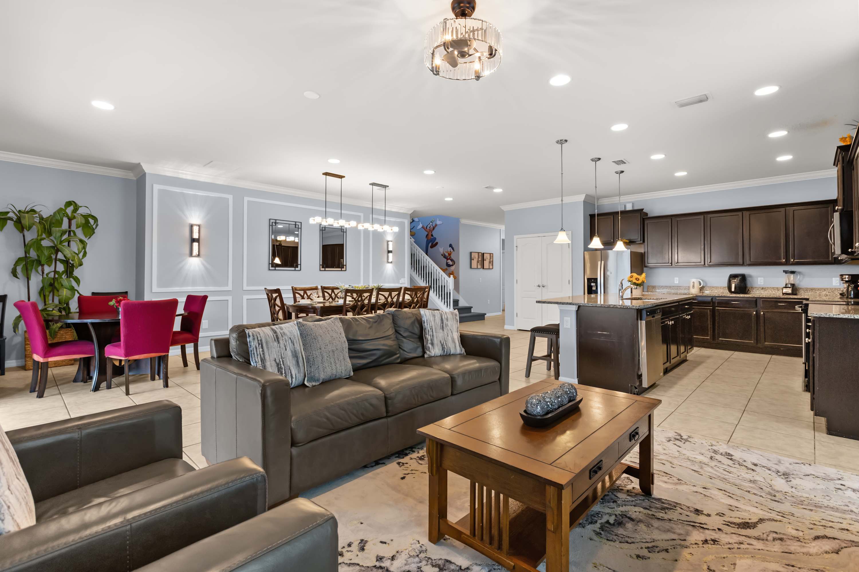 An open-concept living area featuring a dark brown leather sofa and armchairs, a wooden coffee table, and a large patterned rug, flowing into a dining area and kitchen.