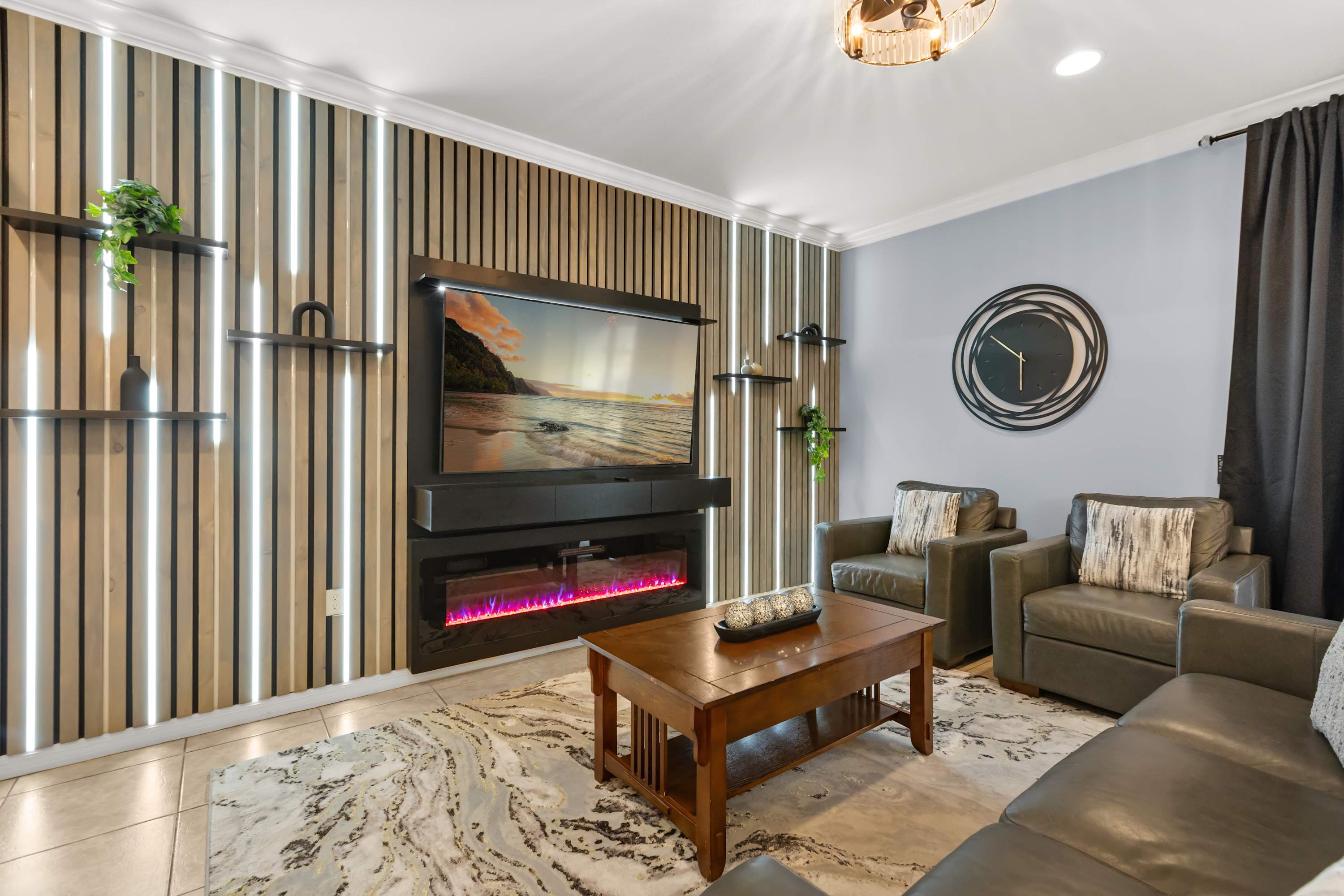 A cozy living room showcases a large flat-screen TV above an electric fireplace, integrated into a stylish slatted accent wall with vertical LED lighting and floating shelves. Two dark leather armchairs and a wooden coffee table are arranged on a patterned rug.