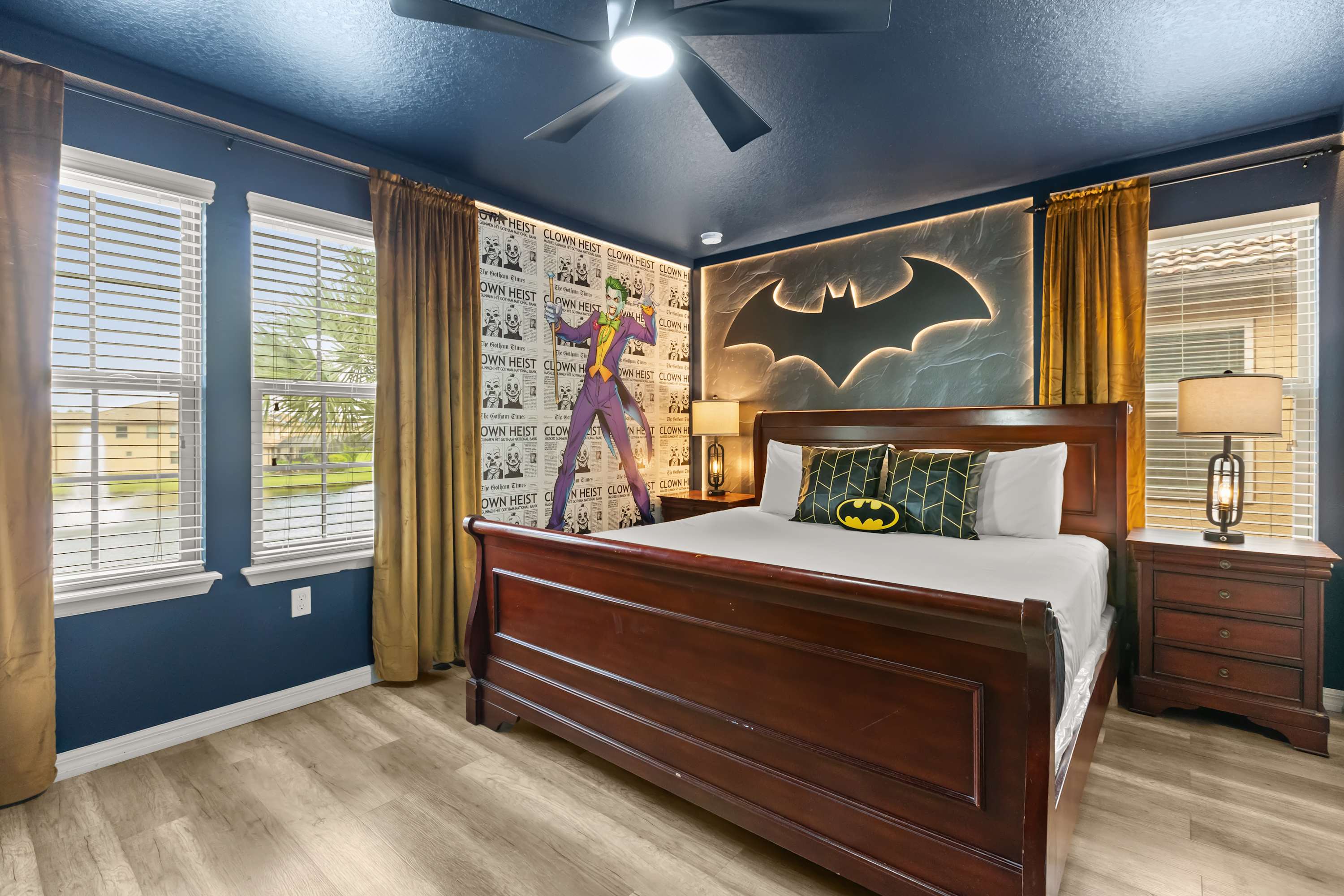 Dark blue Batman-themed bedroom featuring a large wooden bed with a black and yellow pillow, two nightstands with lamps, and a prominent Batman logo wall art with backlighting above the bed. The ceiling is dark blue with a fan.