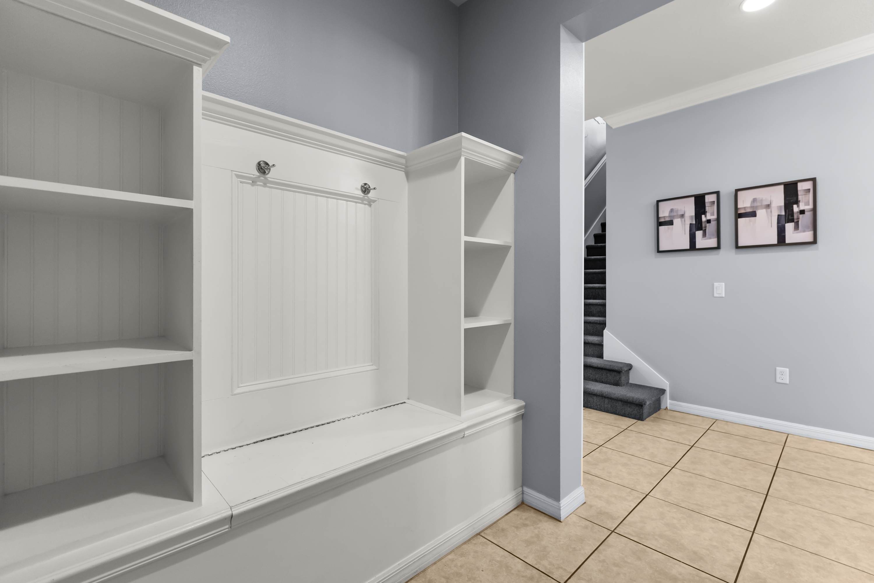 A functional mudroom area features a built-in white bench with hooks and open shelving for storage, providing a practical space near the entrance. The light blue walls and tiled floor maintain a clean and organized aesthetic.