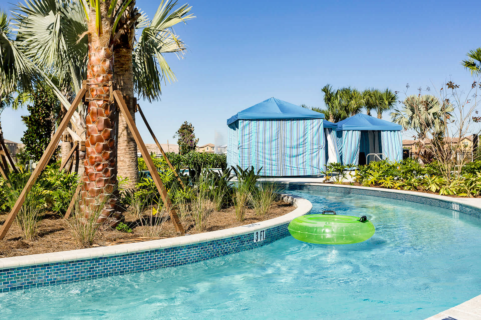 A clear blue lazy river winds through a tropical landscape with palm trees and green foliage. A bright green inflatable tube floats in the water, and blue striped cabanas are visible in the background.