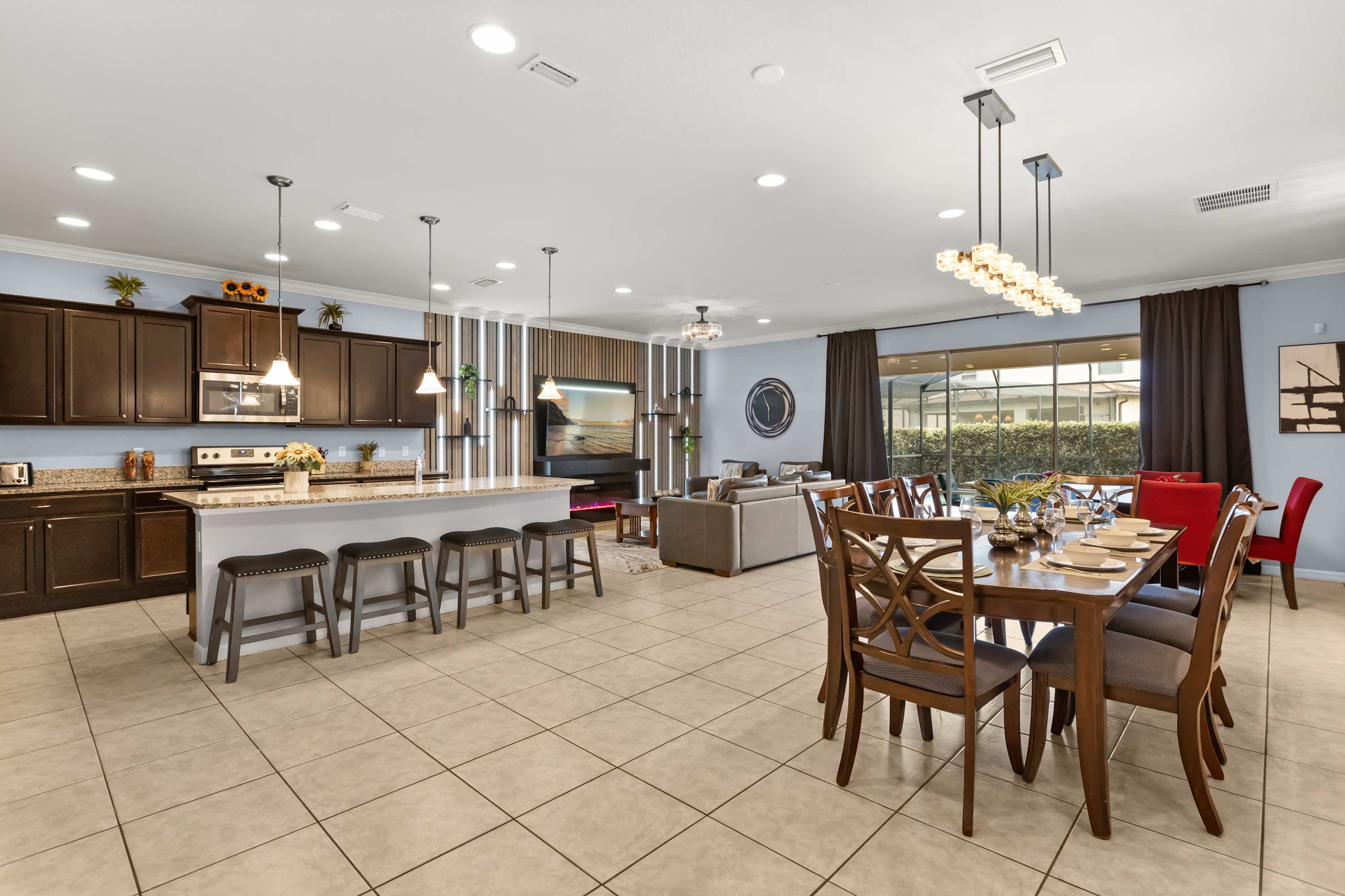 An expansive open-concept living space connects the dining area, kitchen, and living room, featuring a large dining table, a kitchen island with bar stools, and a living area with a TV and fireplace. Tiled floors and light blue walls unify the bright space.