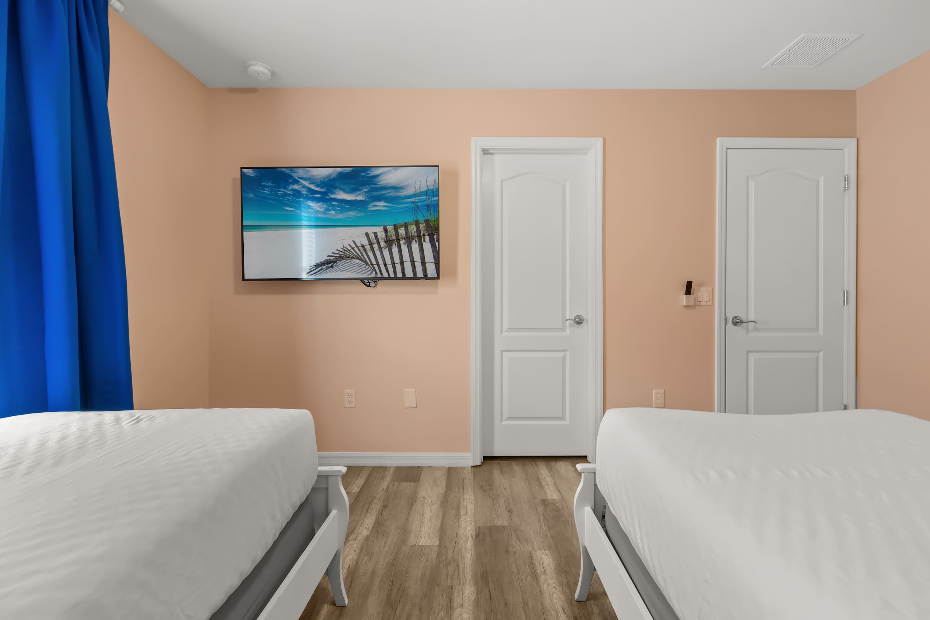 This peach-walled bedroom features light wood-look flooring and a wall-mounted flat-screen TV displaying a beach scene. Two white doors are visible, along with the footboards of two twin beds.