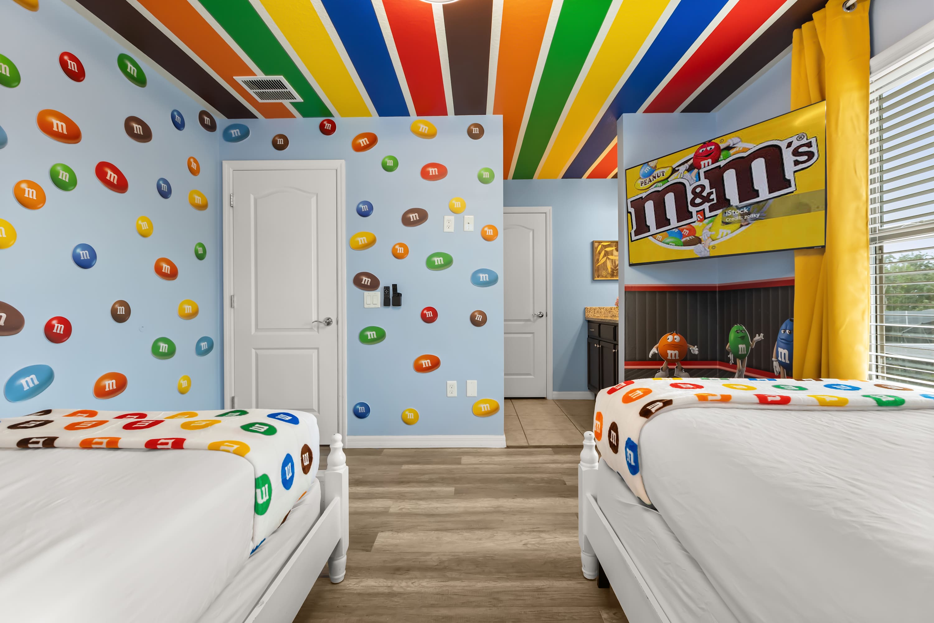 Another perspective of the M&M's themed bedroom, showcasing walls adorned with colorful M&M's decals, two white beds with M&M's themed bedding, and a wall-mounted TV displaying the M&M's logo.