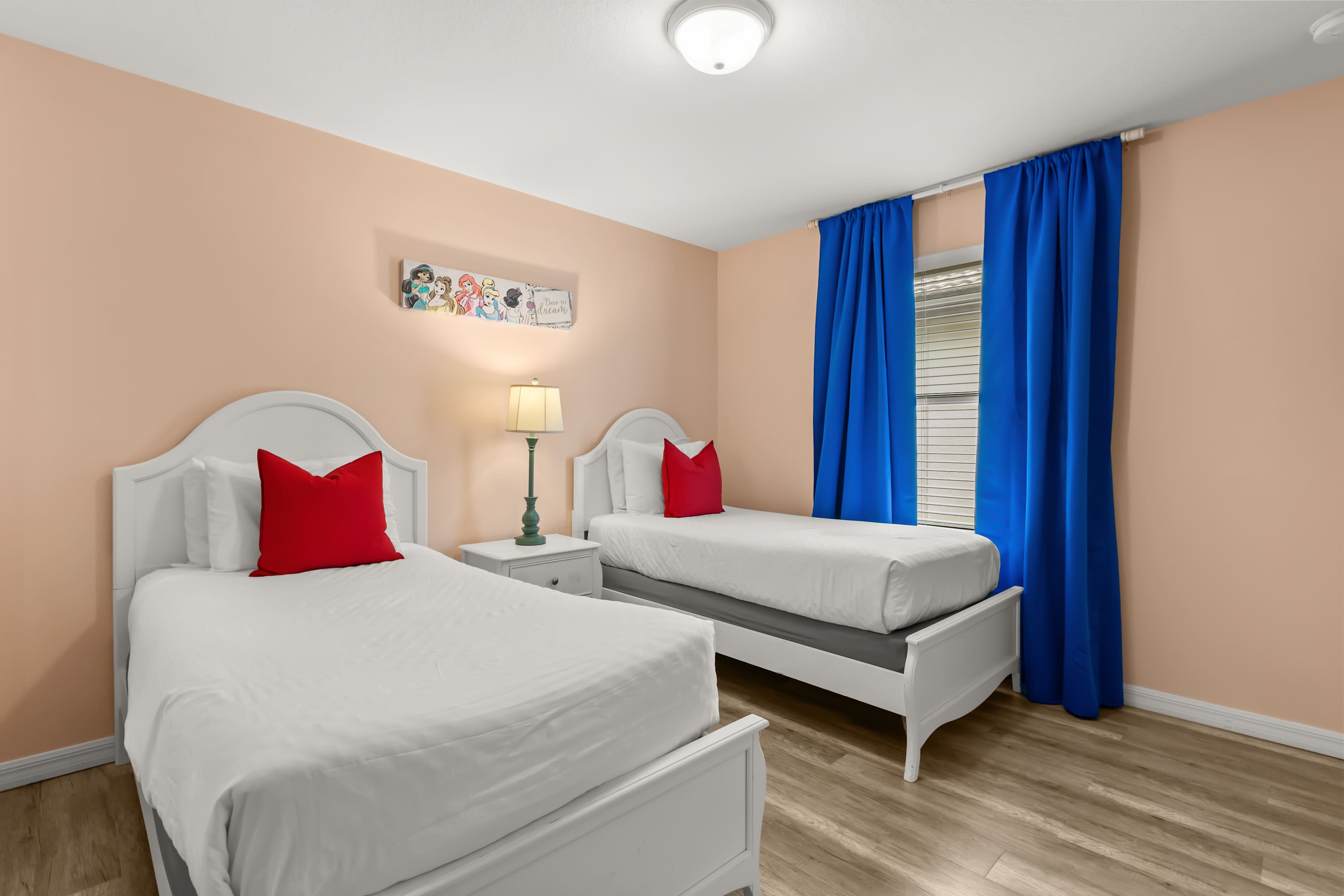 A cheerful bedroom features peach-colored walls, light wood-look flooring, and two white twin beds with white bedding and red accent pillows. A nightstand with a lamp sits between the beds, and a window with blue curtains is visible.
