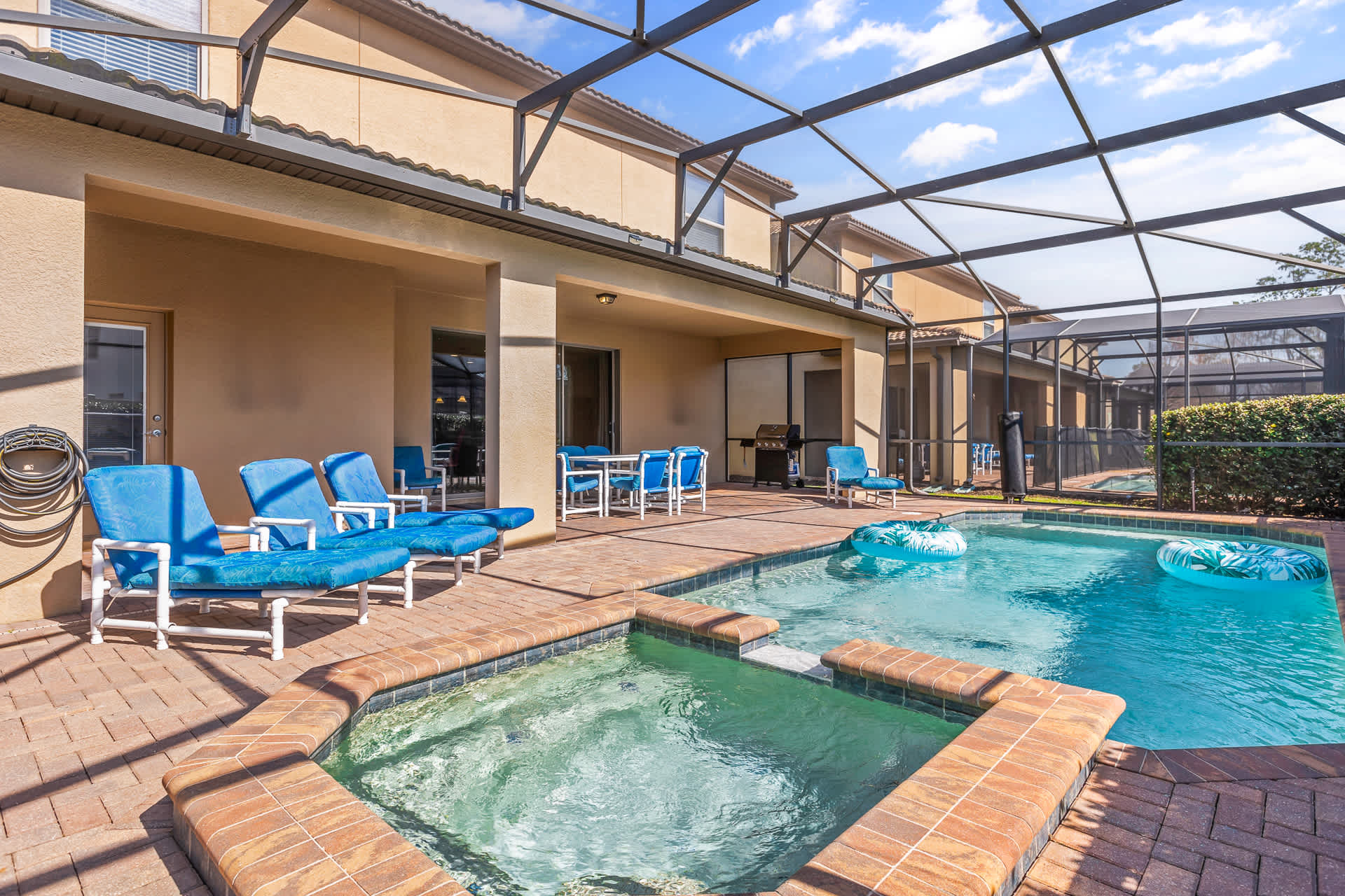 A screened-in private pool area featuring a rectangular pool and an adjacent hot tub, surrounded by a brick patio with blue lounge chairs and an outdoor dining set.
