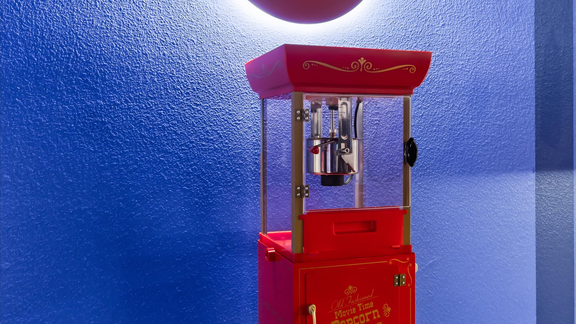 A close-up view of a classic red and gold popcorn machine, featuring a glass enclosure and "Old Fashioned Movie Time Popcorn" text, set against a vibrant blue wall.