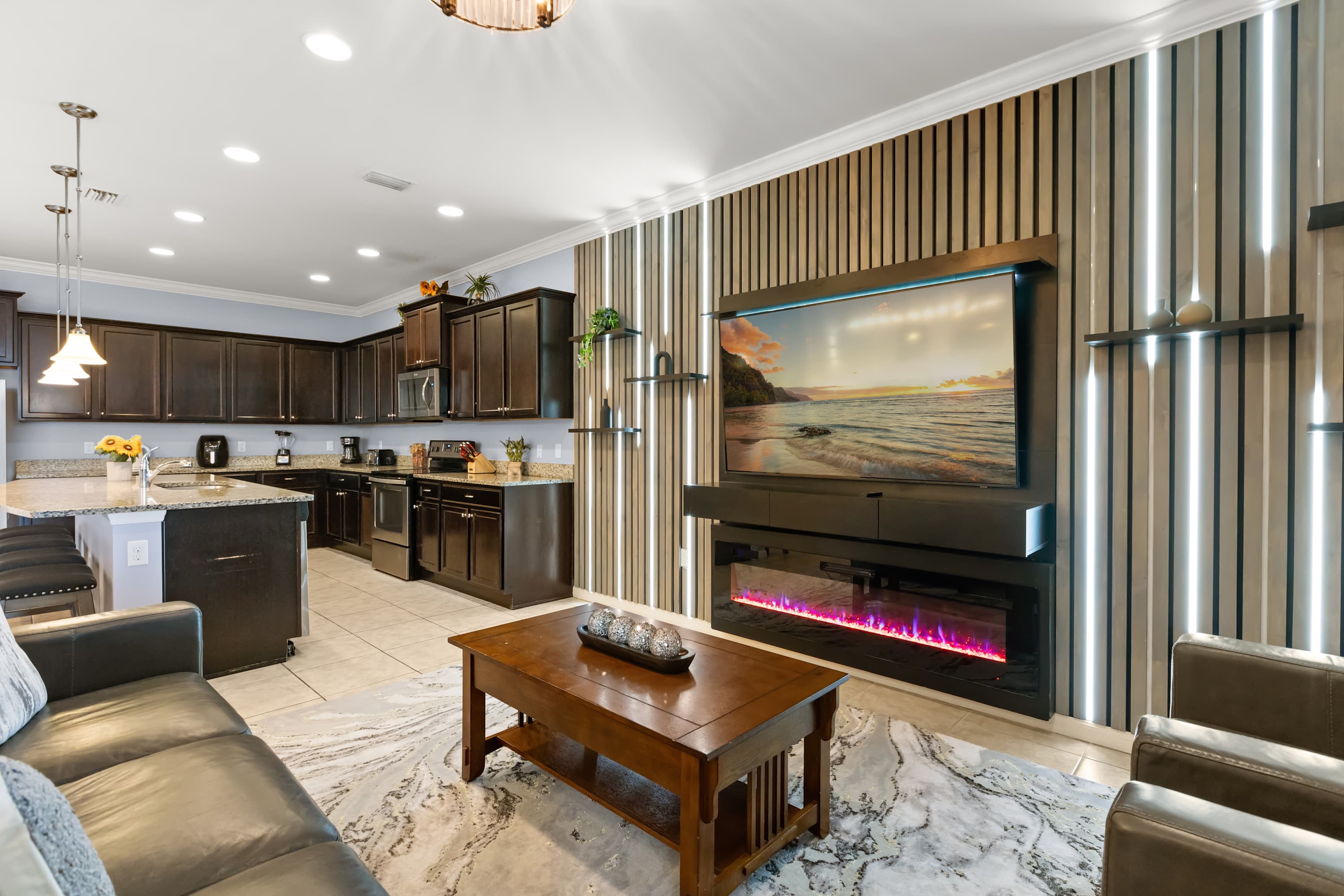 An open-concept living area features a large flat-screen TV mounted above an electric fireplace, set against a modern slatted accent wall with vertical lighting. A dark leather sofa and armchair face a wooden coffee table, with a kitchen visible in the background.
