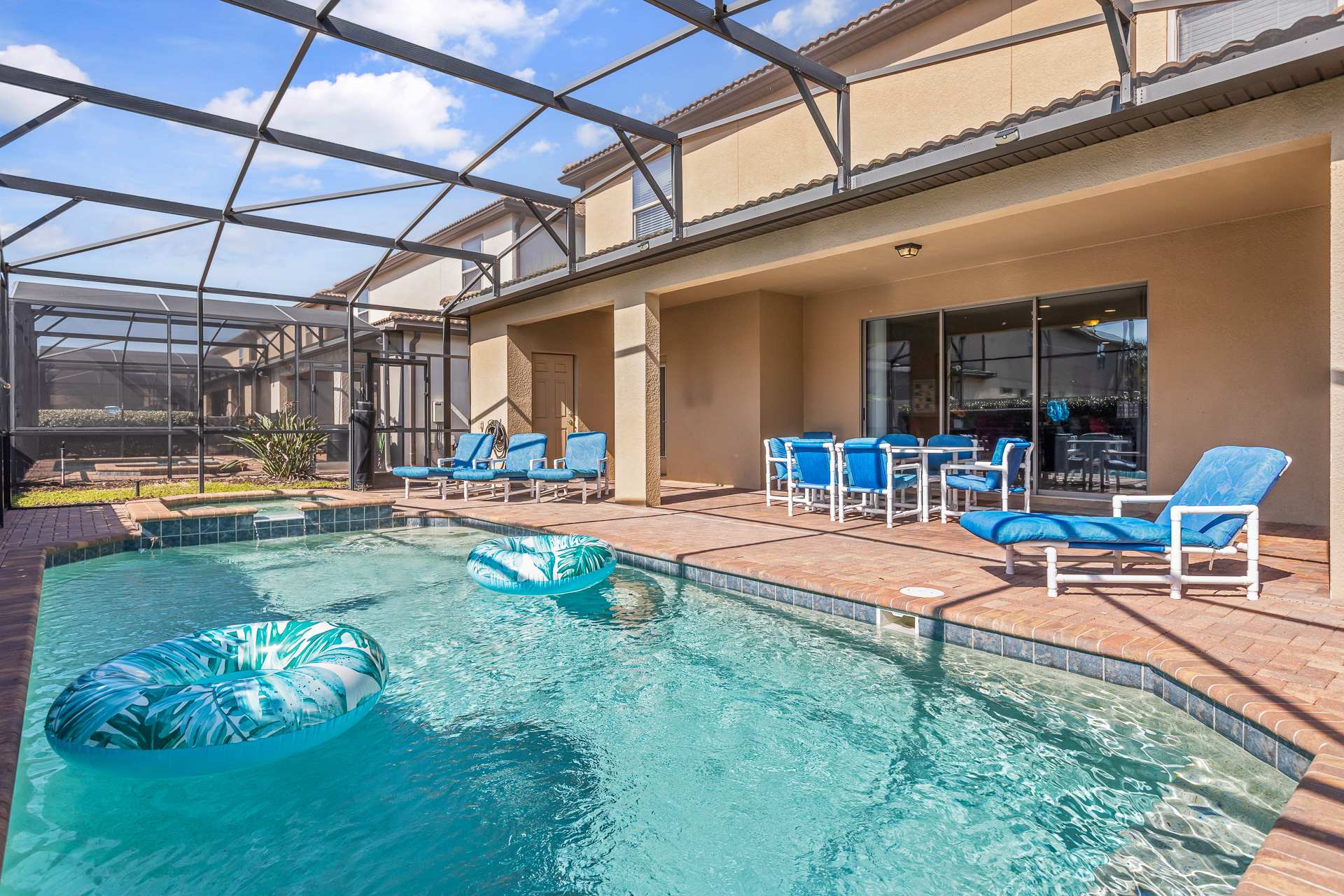 A screened-in private pool area with a rectangular pool, several blue lounge chairs, and an outdoor dining table with blue chairs under a covered patio.