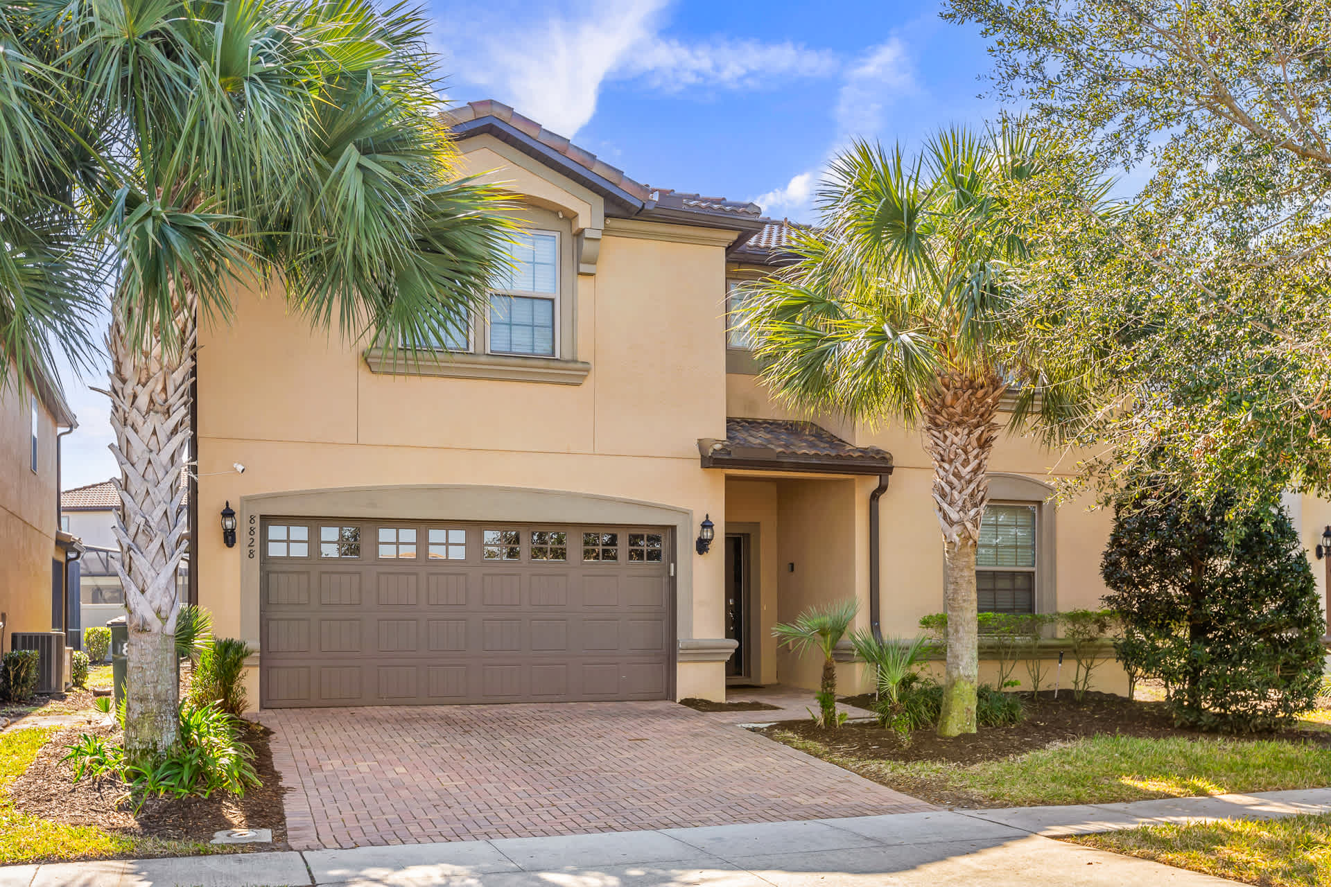 The front exterior of a two-story house with a light tan stucco facade and a brown tile roof. It features a two-car garage with a brick paver driveway, surrounded by palm trees and lush landscaping under a blue sky.