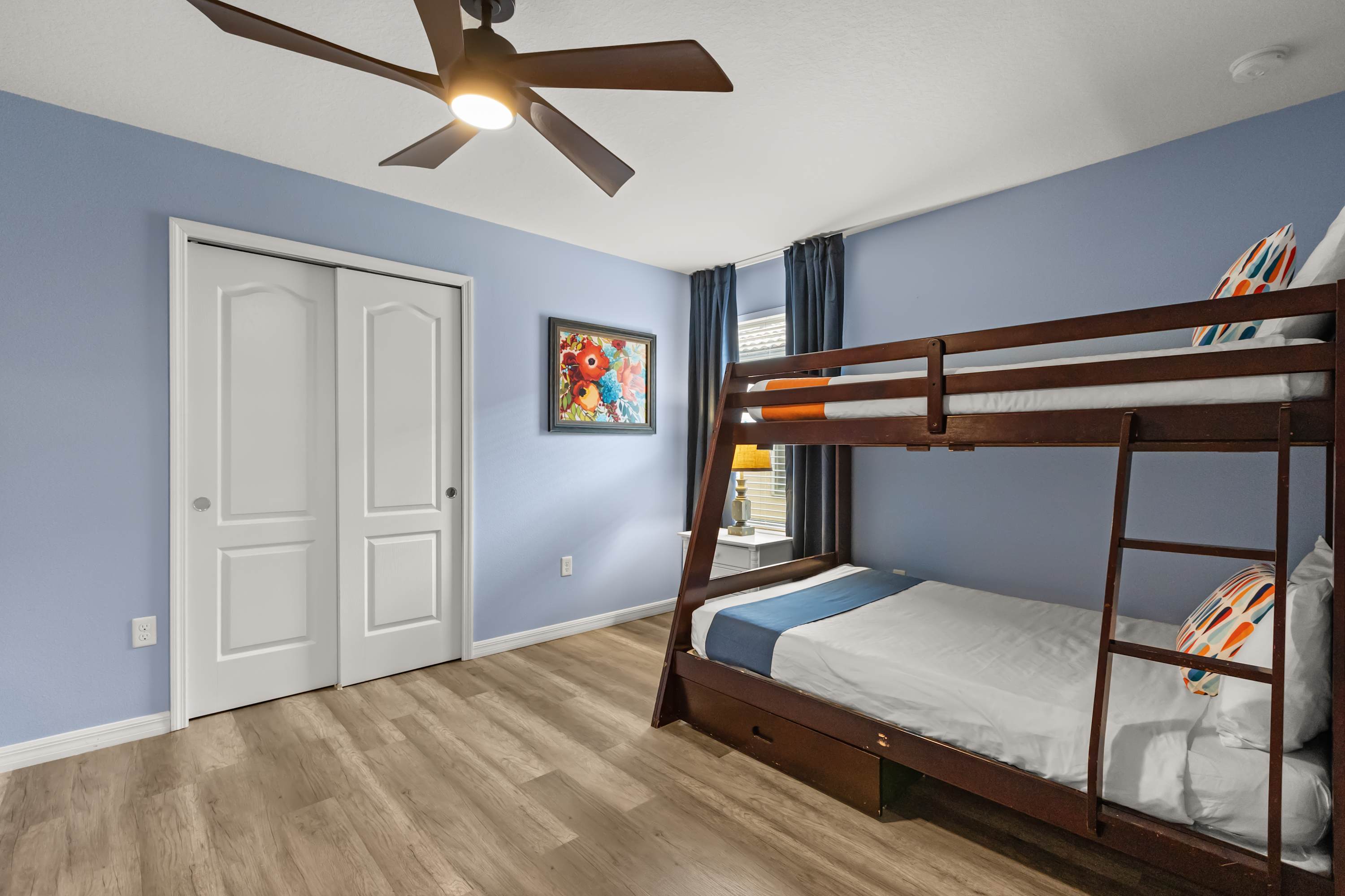 A vibrant bedroom features light blue walls, light wood-look flooring, and a dark wooden bunk bed with white bedding. A ceiling fan with integrated light fixture hangs overhead, and a colorful floral painting adorns the wall.