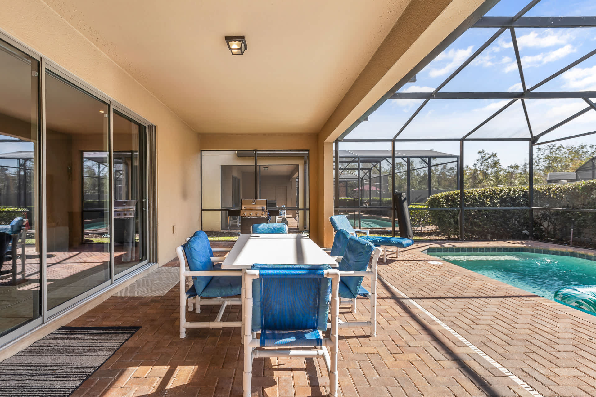 A covered outdoor patio area with a white dining table and blue chairs, overlooking a screened private pool, with sliding glass doors leading into the house.