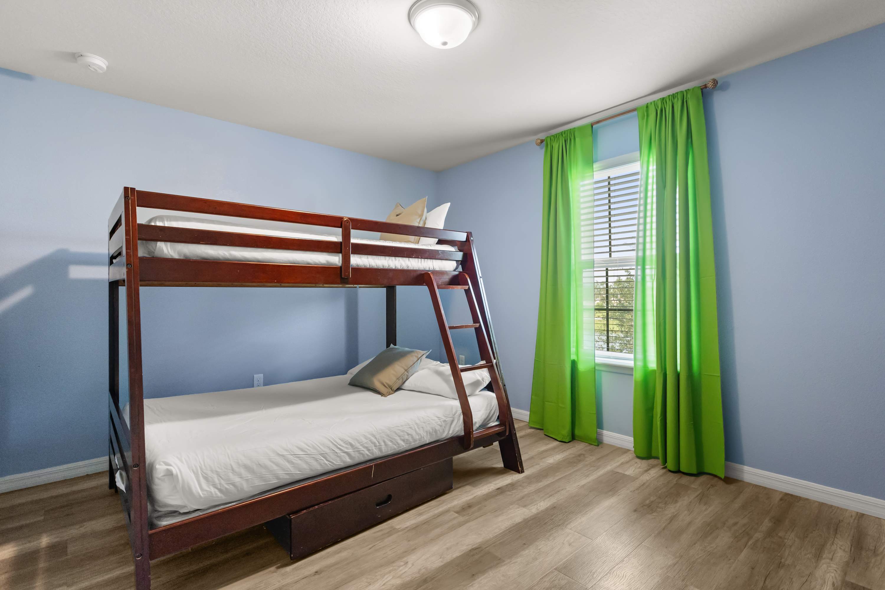 A bright bedroom features light blue walls, light wood-look flooring, and a dark wooden bunk bed with white bedding. A window with white blinds is framed by vibrant lime green curtains, and a flush-mount ceiling light illuminates the room.