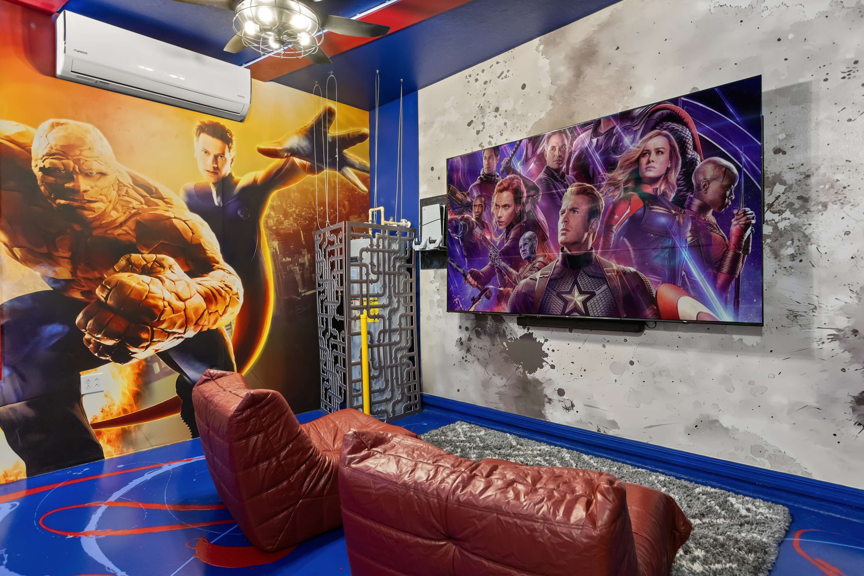A themed game room with a large TV displaying Marvel characters, two red bean bag chairs, and a vibrant mural featuring The Thing and other superheroes on the walls.