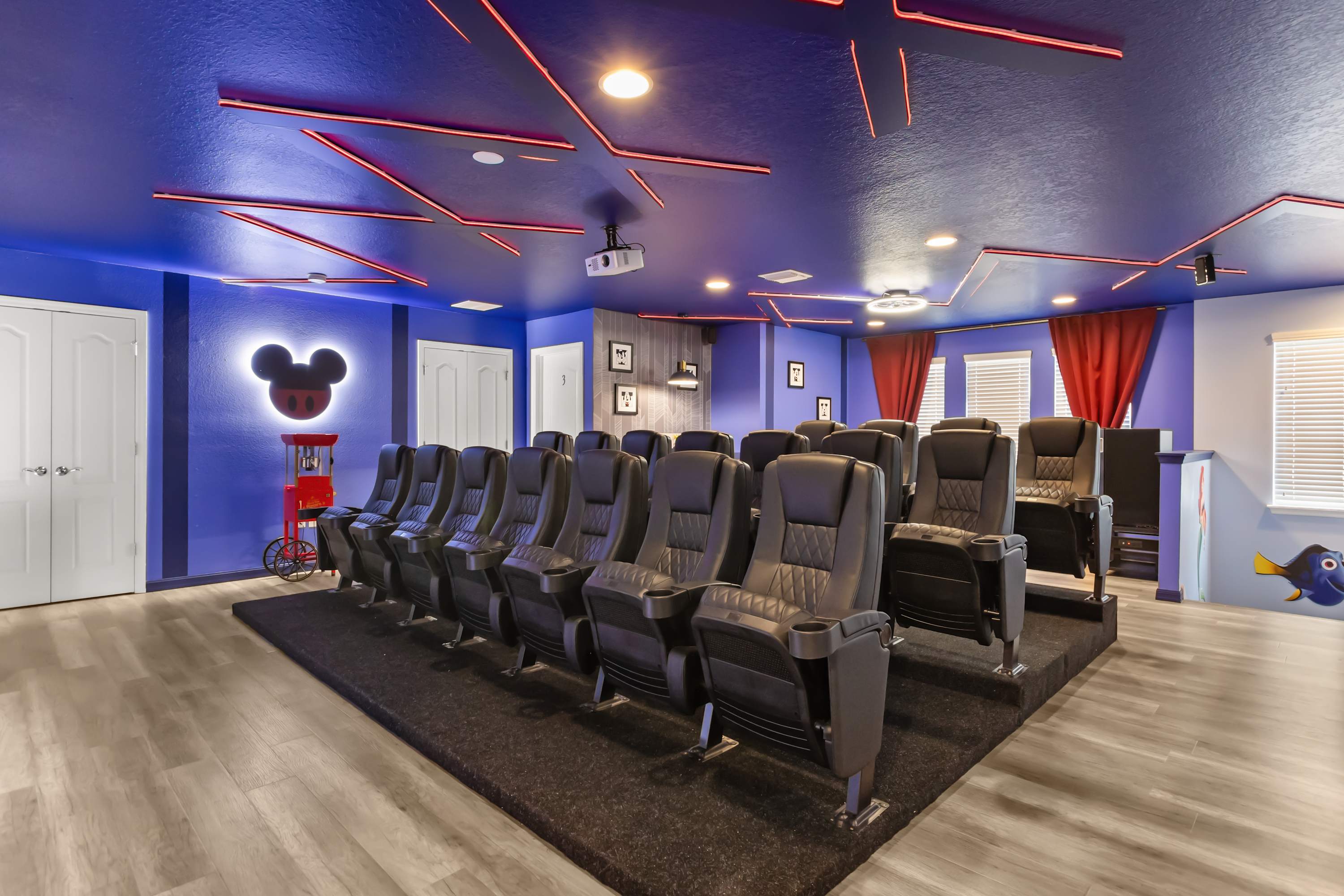 A dramatic home theater features deep blue walls and ceiling with red accent lighting, tiered rows of black leather recliners, and a projector. A Mickey Mouse silhouette light and a popcorn machine add to the themed decor.