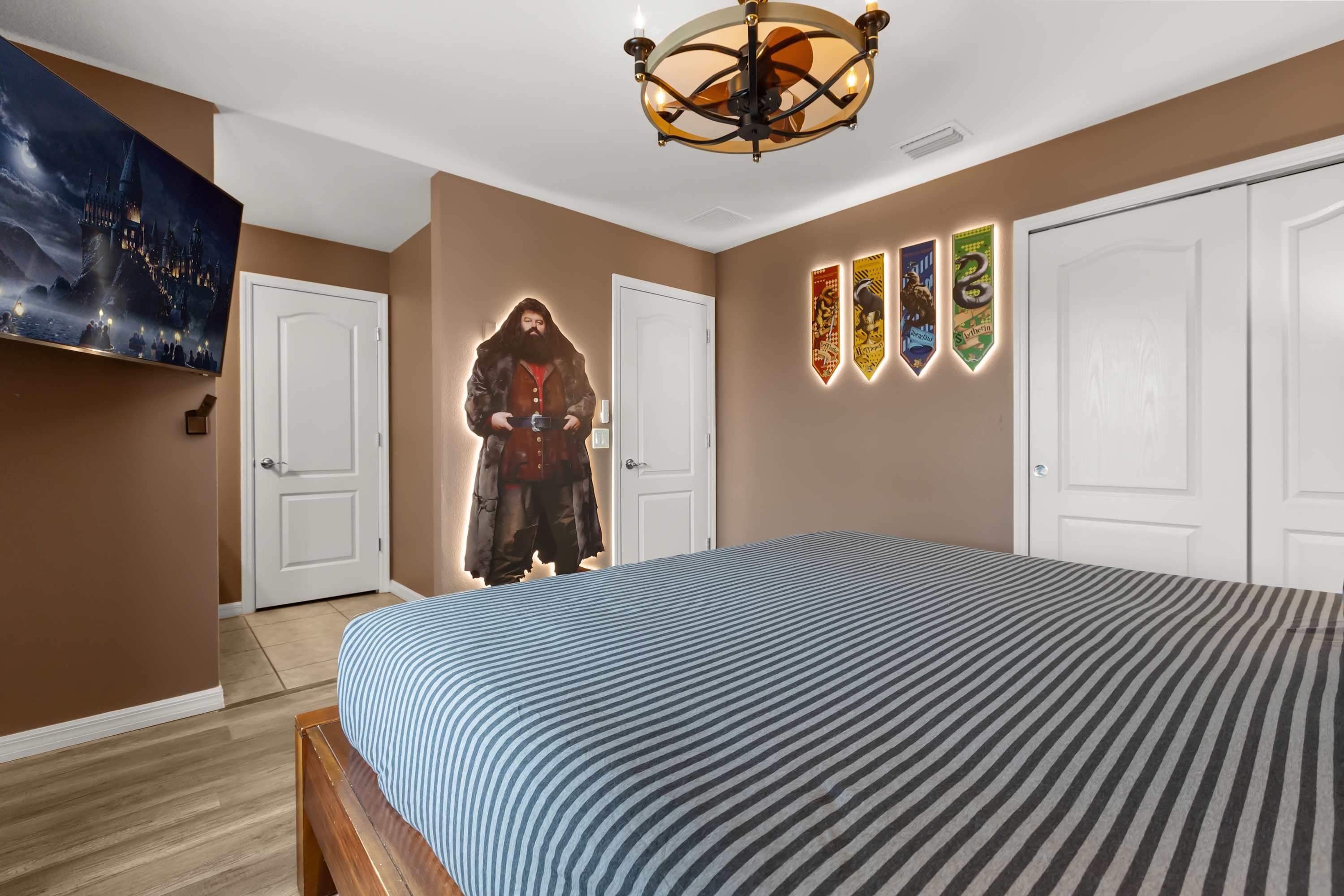 Themed bedroom with a large bed featuring a striped duvet, a wall-mounted TV displaying Hogwarts, and a life-size Hagrid cutout with backlighting. The walls are painted a warm brown, and there's a ceiling fan with a light fixture.