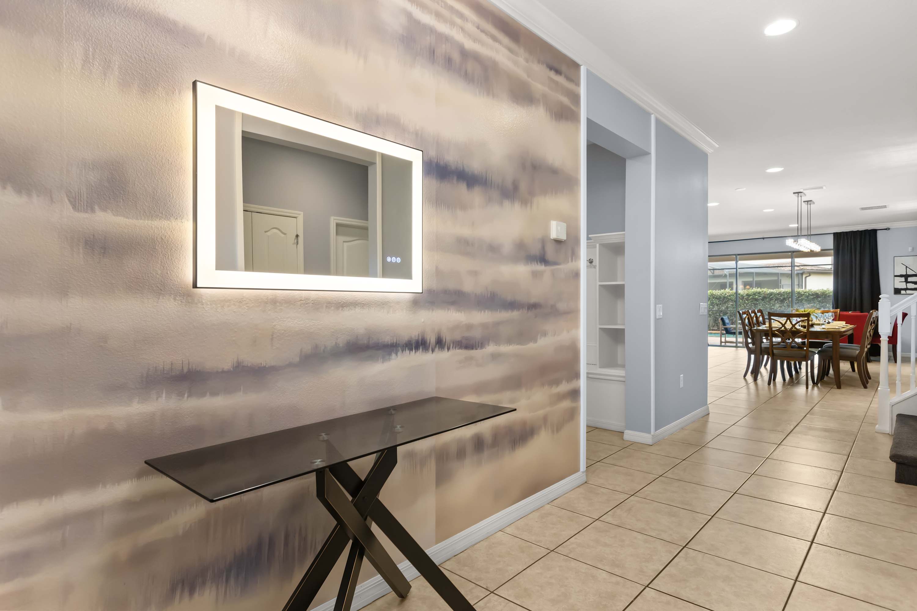 A welcoming foyer features a modern console table with a black base and a large rectangular illuminated mirror, set against a textured accent wall with abstract horizontal brushstroke patterns in muted tones. Tiled flooring extends into the adjacent hallway.