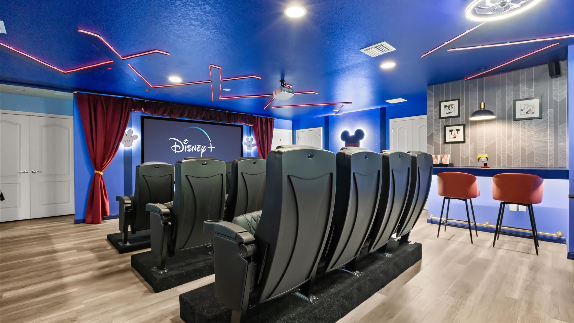 A home theater room with a large screen displaying Disney+, multiple rows of black cinema seats, a blue ceiling with red neon lighting, and a small bar area with red stools.