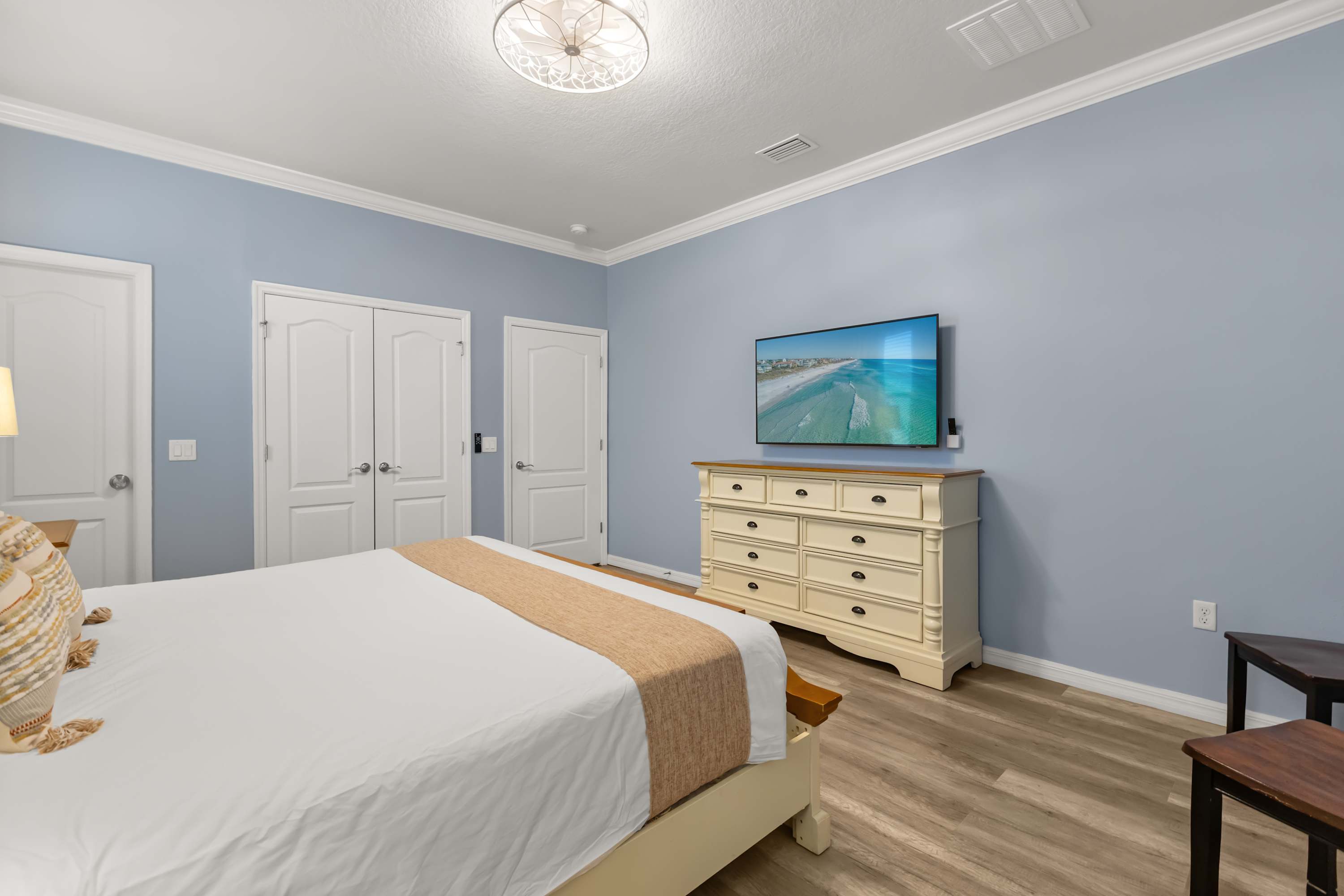 A bright bedroom features light blue walls and light wood-look flooring, furnished with a cream-colored bed and a matching dresser with a flat-screen TV mounted above it. White double doors and a decorative ceiling light complete the serene space.