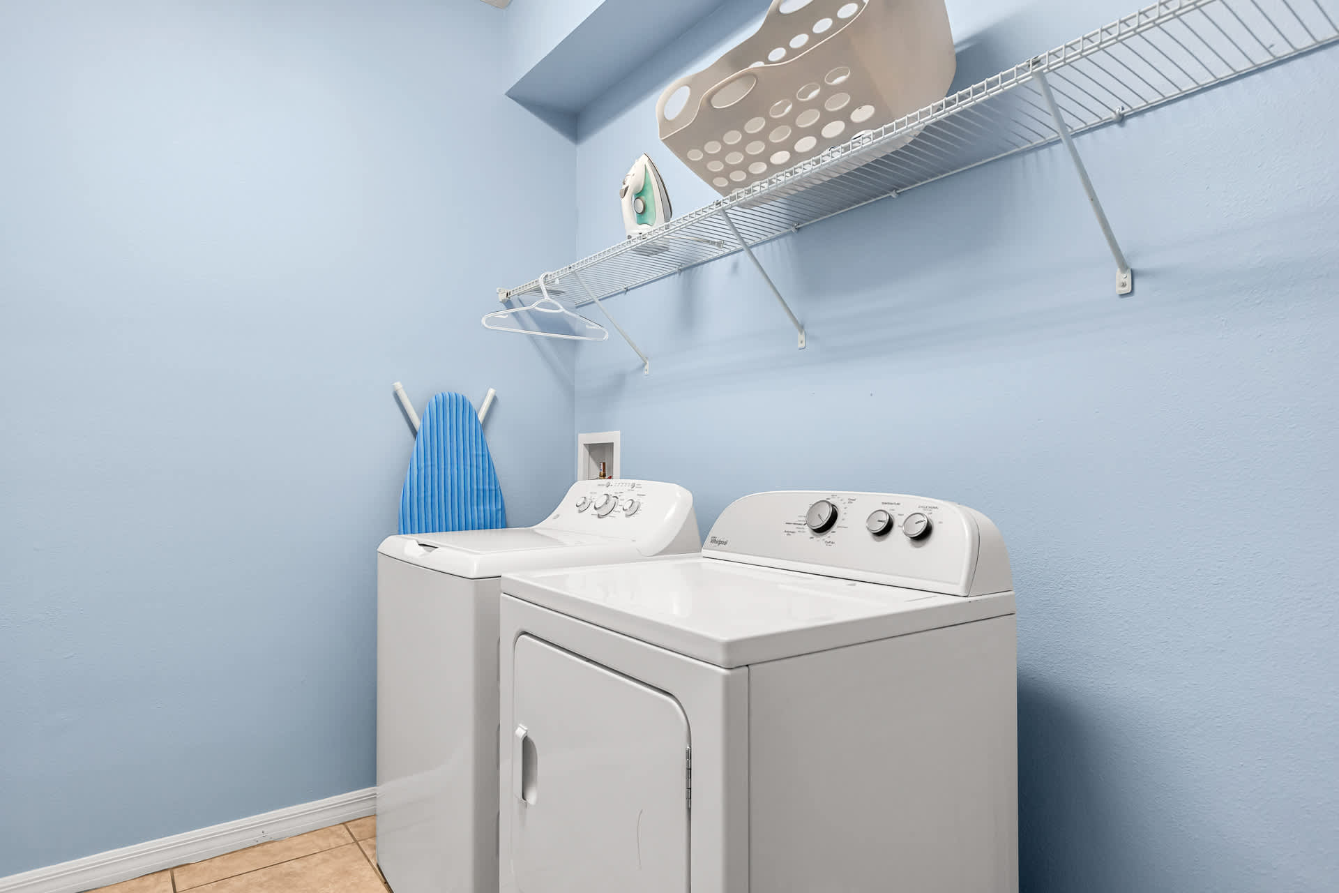 A bright laundry room with light blue walls features a white top-loading washing machine and a front-loading dryer. A wire shelf above holds a laundry basket and iron, with an ironing board leaning against the wall.