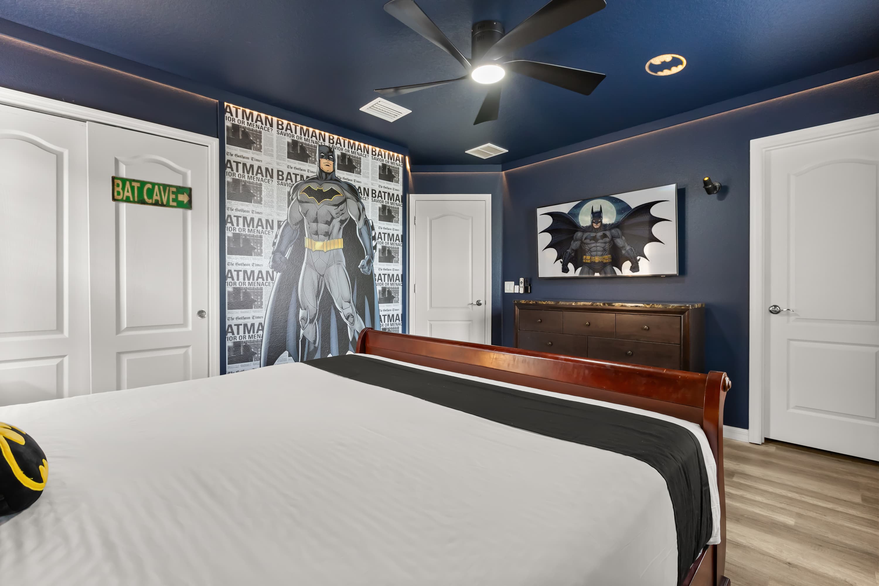 Another view of the Batman-themed bedroom, showcasing a large Batman cutout against a patterned wall, a "BAT CAVE" sign on a white door, and a wall-mounted TV displaying Batman art. A dark wooden dresser is also visible.