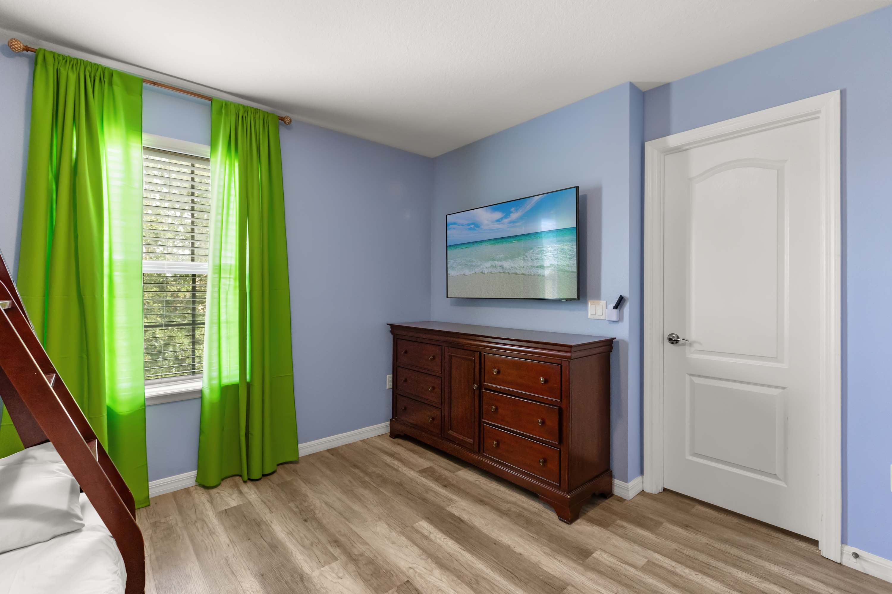 This light blue bedroom features light wood-look flooring, a dark wooden dresser, and a wall-mounted flat-screen TV displaying a beach scene. A window with green curtains and a white door are also visible.
