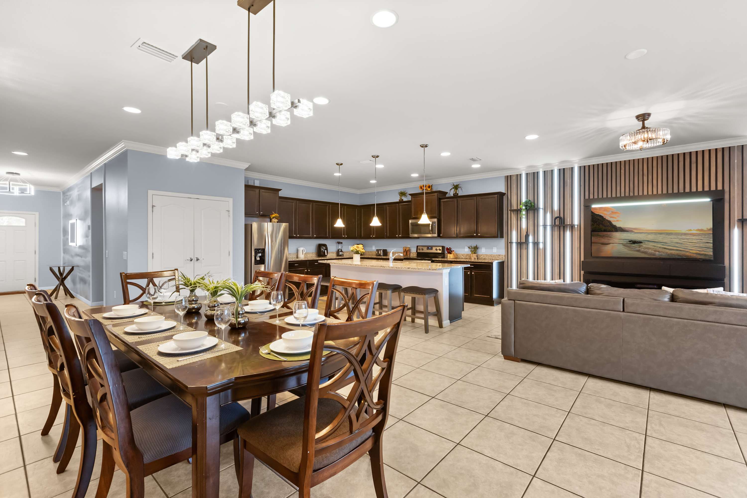 A spacious open-concept area combines a formal dining space with a view of the kitchen and living room, featuring a large dark wood dining table with eight chairs under a modern linear chandelier. Light blue walls and tiled floors create a bright atmosphere.