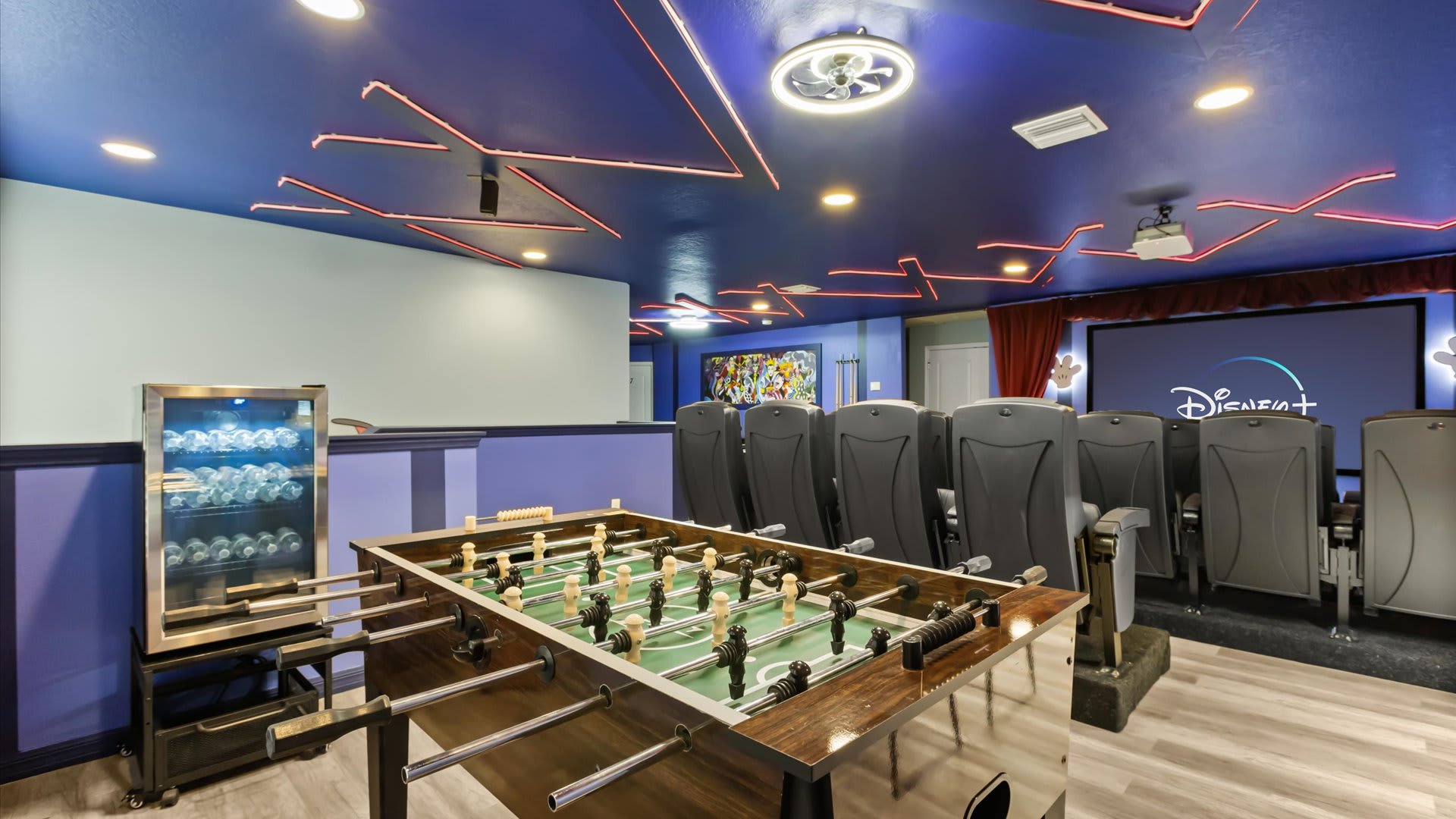 A vibrant media room features a foosball table in the foreground, a beverage cooler, and a large projection screen displaying Disney+. The room has a blue ceiling with red neon strip lighting and stadium seating.