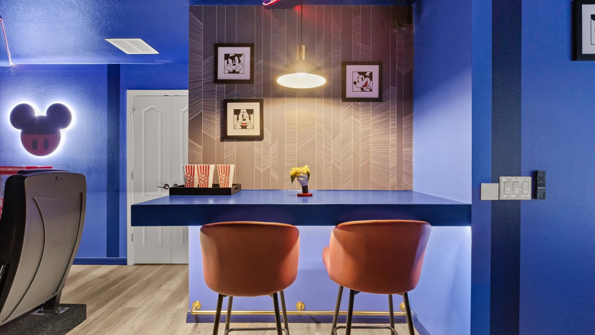 A themed bar area features deep blue walls, a blue countertop with underlighting, and two orange bar stools. Mickey Mouse framed artwork and a hanging pendant light complete the space, with a popcorn machine visible nearby.