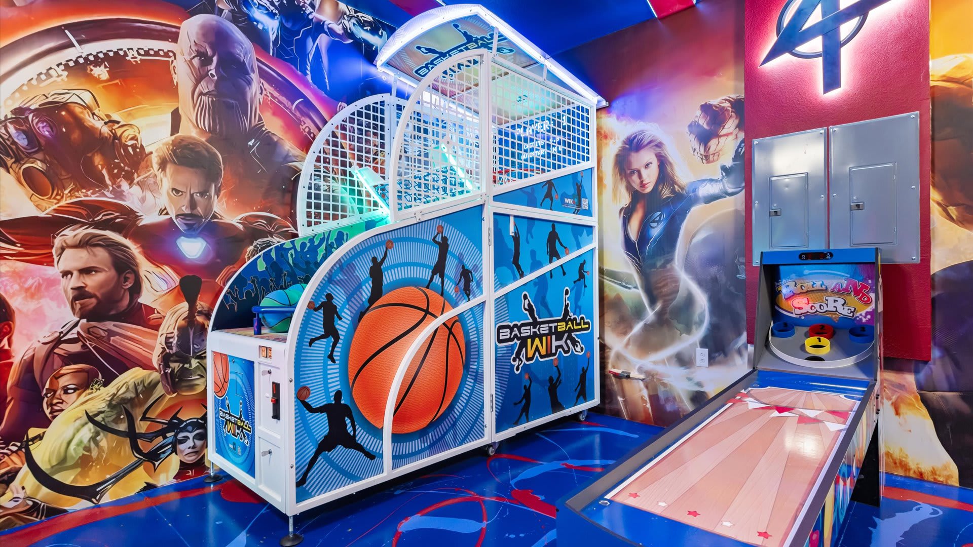 A vibrant game room features an arcade basketball game and a skee-ball machine, set against walls adorned with colorful superhero murals and illuminated by blue and white lighting. The floor is a dark blue with red and white abstract patterns.