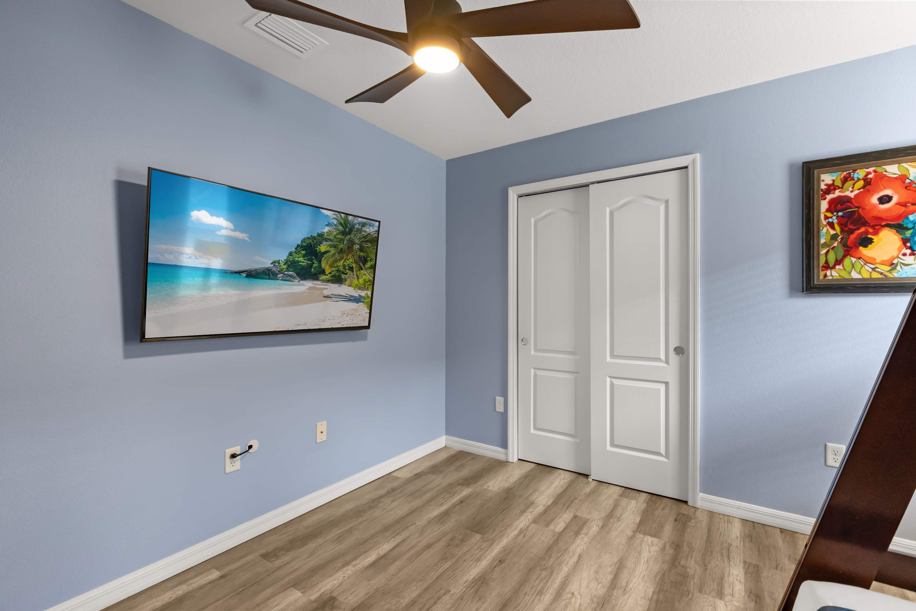 This light blue bedroom features light wood-look flooring and a large wall-mounted flat-screen TV displaying a beach scene. A white bi-fold closet door is visible, and a ceiling fan is partially seen overhead.