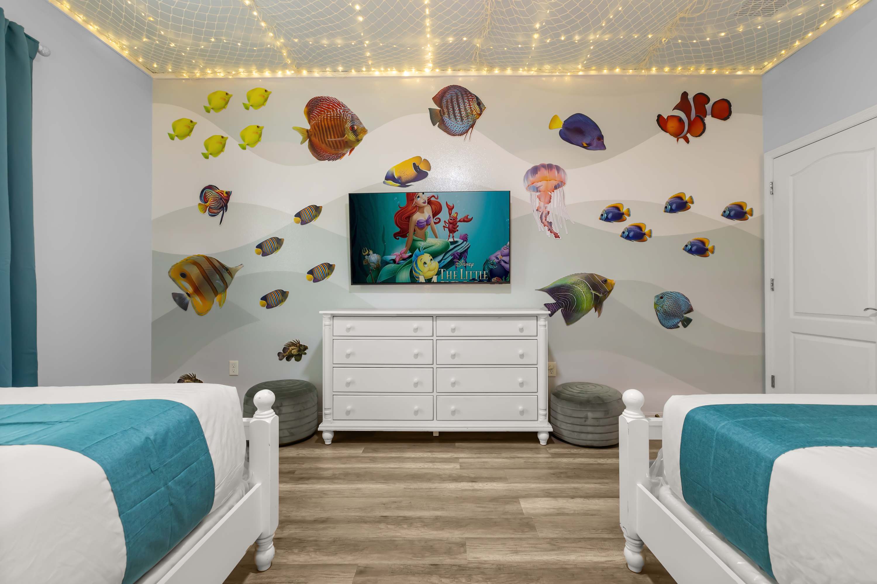 Another view of the ocean-themed bedroom, featuring two white beds, a white dresser with a wall-mounted TV displaying "The Little Mermaid," and various fish decals on the light blue wall.